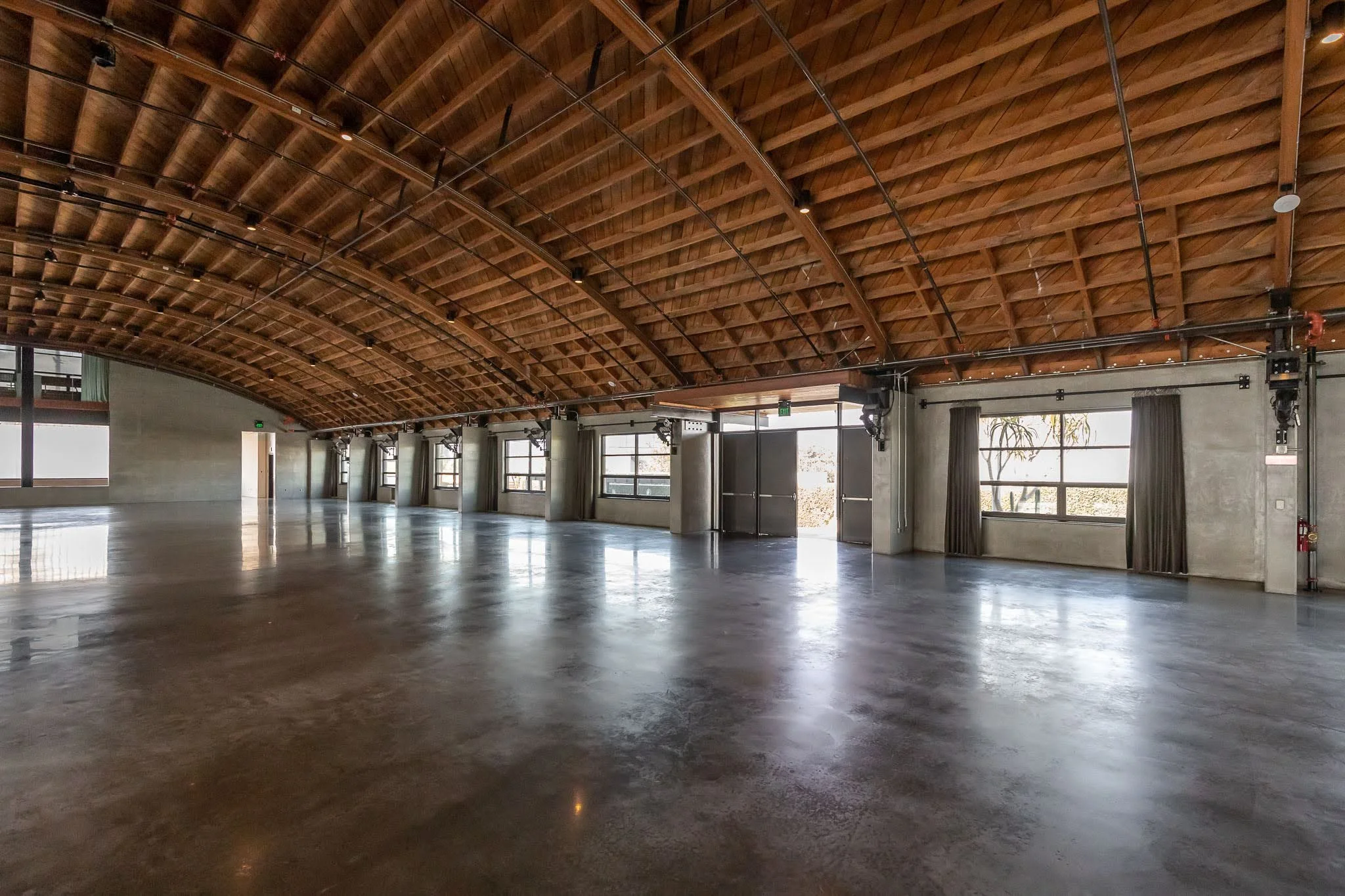 Empty spacious event hall with polished concrete floor, large windows with curtains, and a curved wooden ceiling.