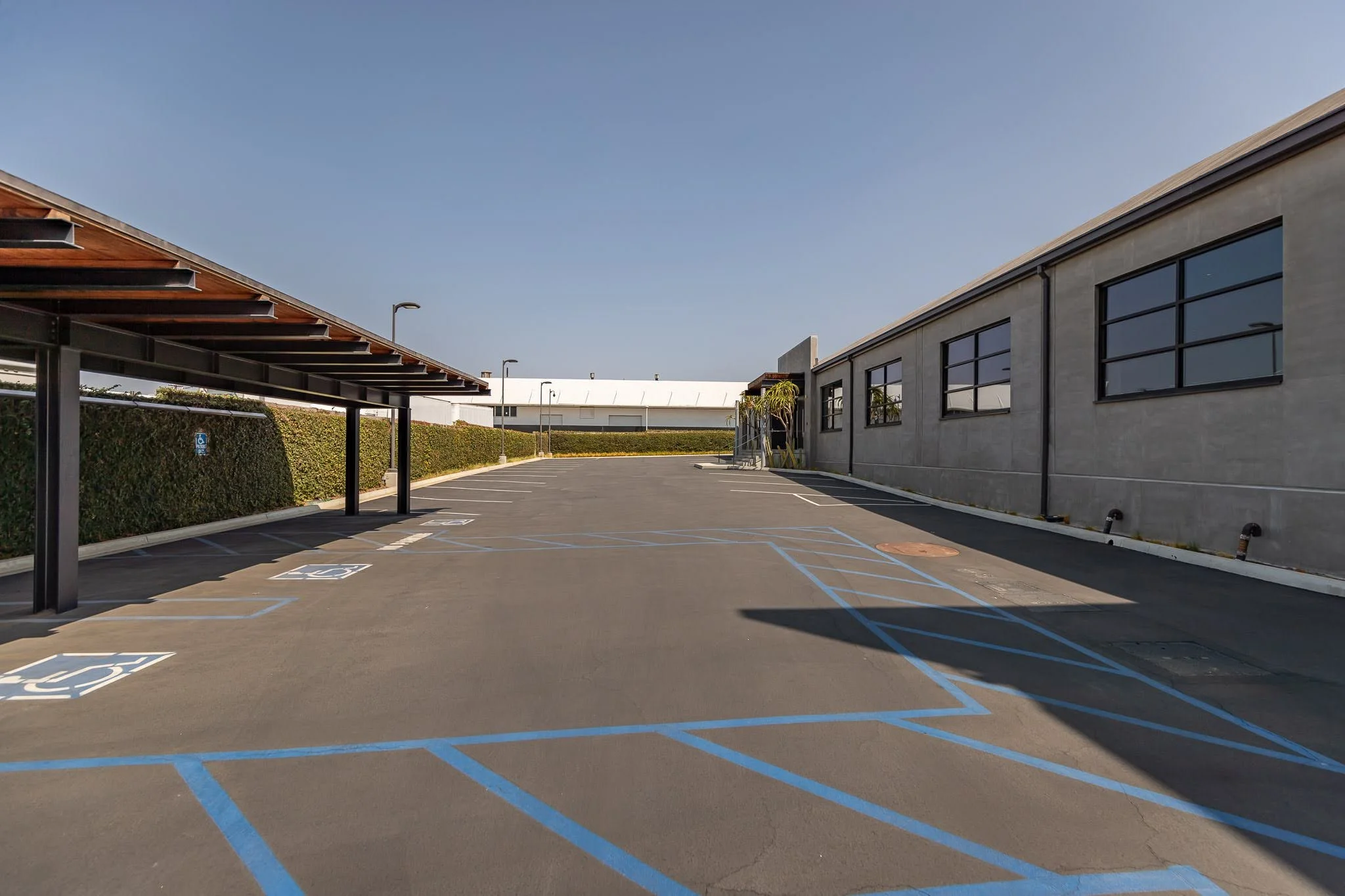 Empty parking lot with designated handicap parking spaces and a concrete building on the right, hedge along the back, and a clear blue sky overhead.