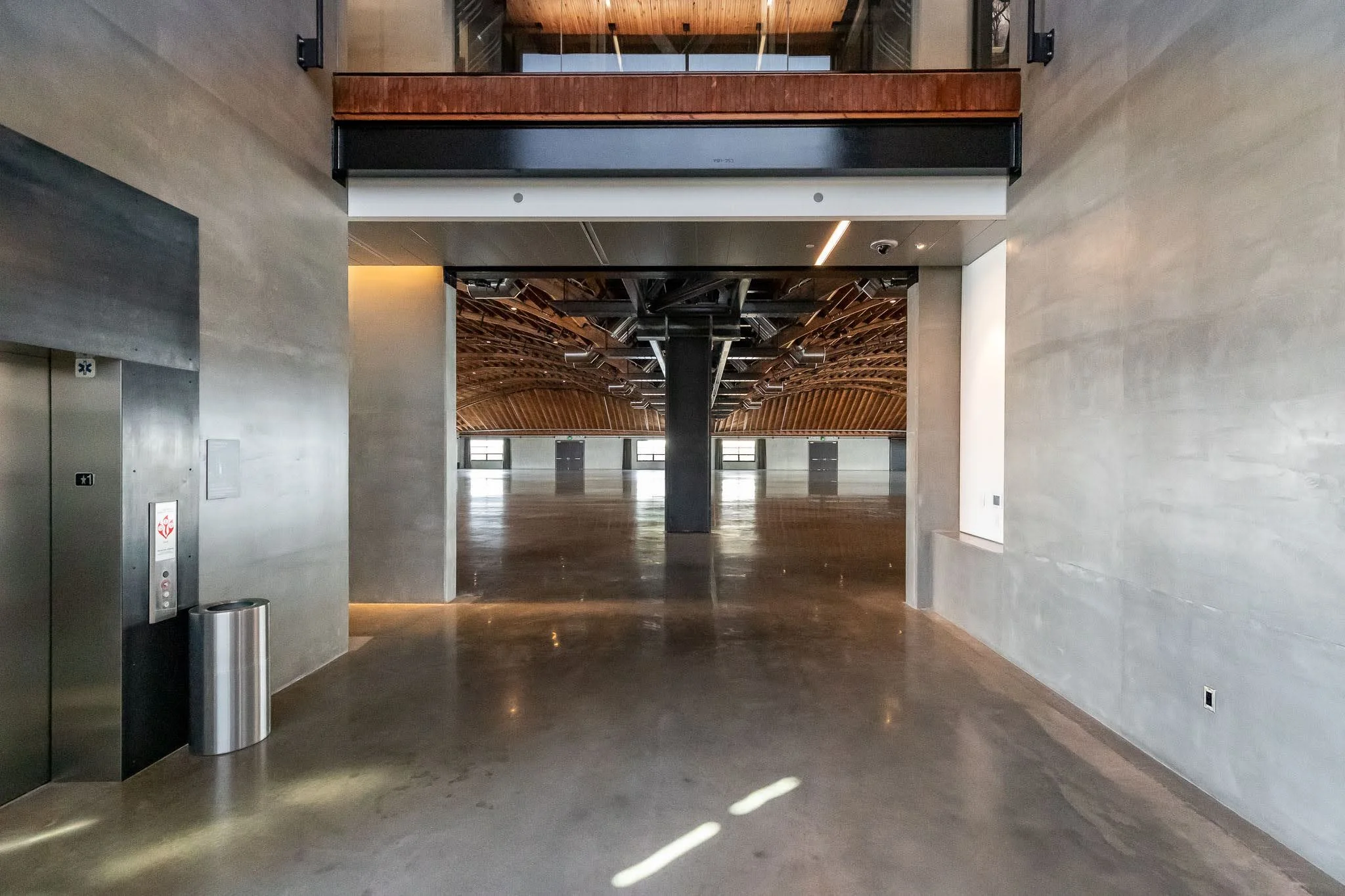 Interior view of a large, empty, modern space with polished concrete floors, concrete walls, and a curved wooden ceiling in the background. An elevator is on the left side of the image.