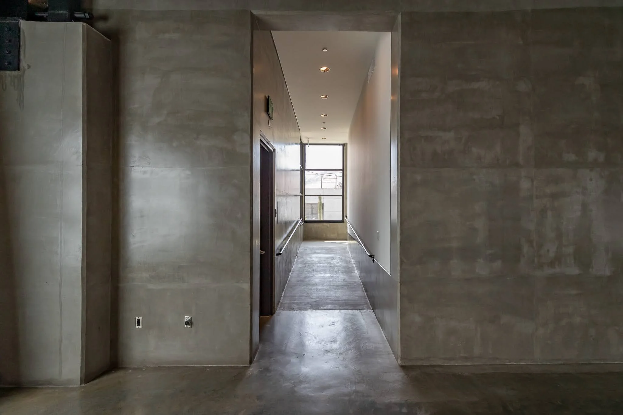 Concrete hallway with window at the end, three ceiling lights, and electrical outlets on the wall.