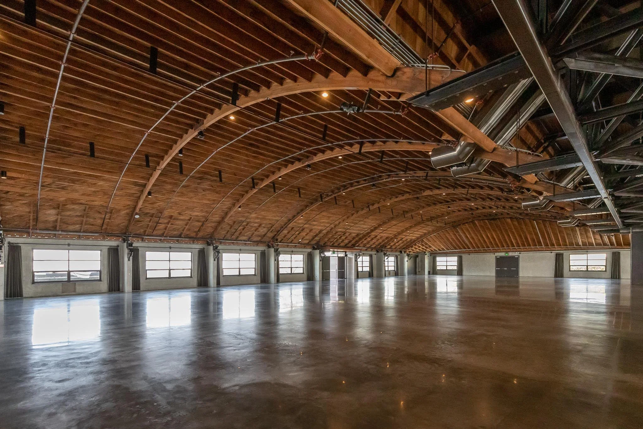 Interior of a spacious, empty hall with a polished concrete floor, wooden arched ceiling, and multiple windows along the walls.