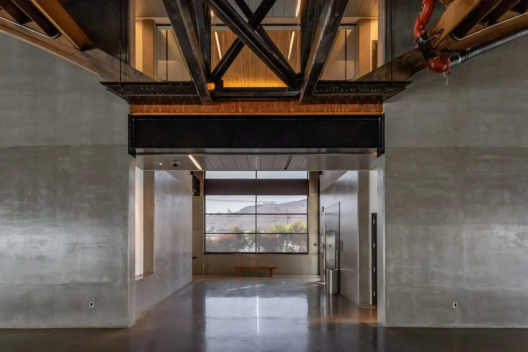 Interior view of a modern building with a concrete wall, large window, and polished floor, featuring exposed black steel beams and a small bench near the window.