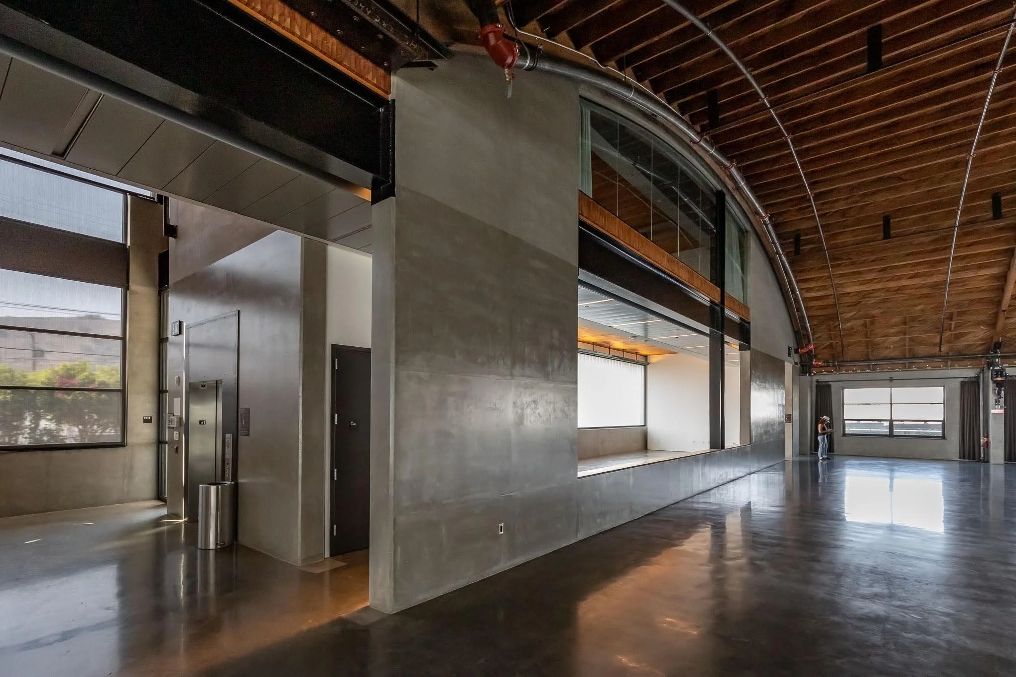 Interior of a modern, spacious building with concrete walls, large windows, and a wooden arched ceiling. There is an elevator on the left and a person near the far window on the right.