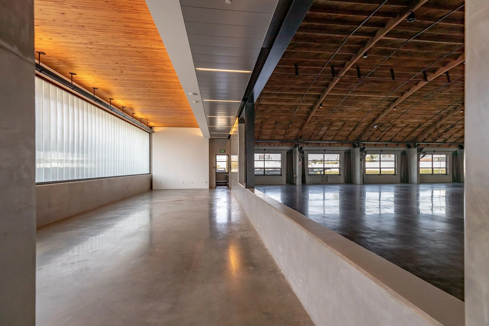 Interior of a spacious, modern building with a polished concrete floor, large windows, wooden and black metal ceiling, and a separated open area with tall ceilings and multiple windows.