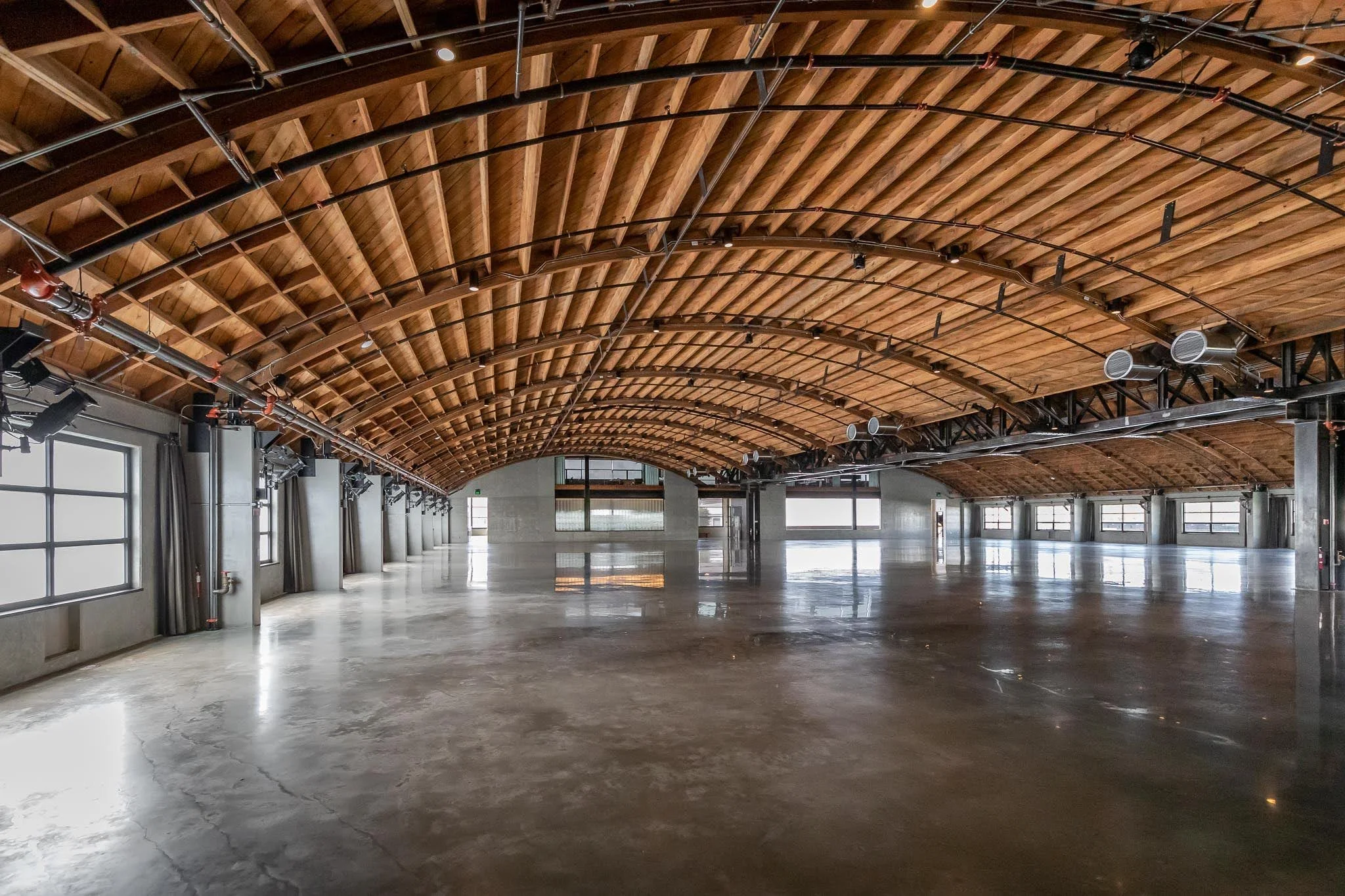 Spacious empty indoor space with polished concrete floor, wood arched ceiling, large windows, and industrial lighting.
