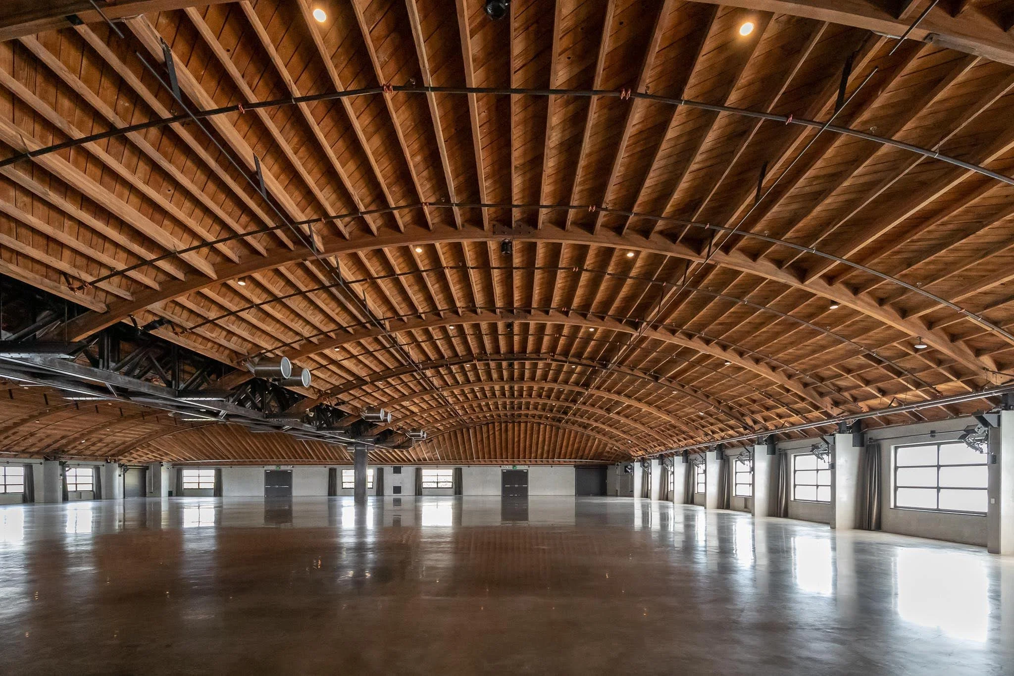 Empty large room or event space with polished concrete floor, curved wooden ceiling, and multiple large windows along the walls.