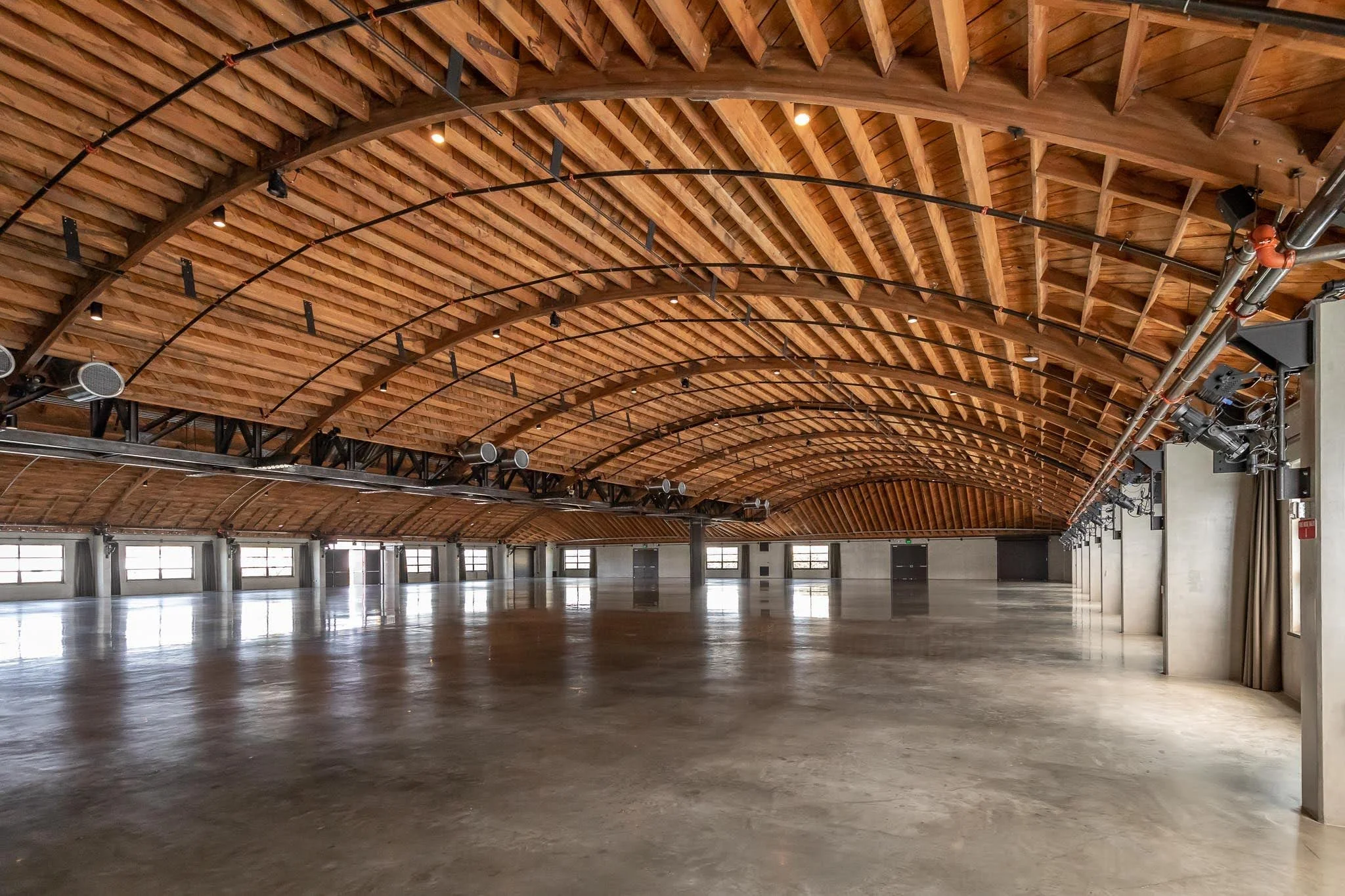 Large empty event hall with polished concrete floor, wooden arched ceiling, and multiple windows along the side walls.