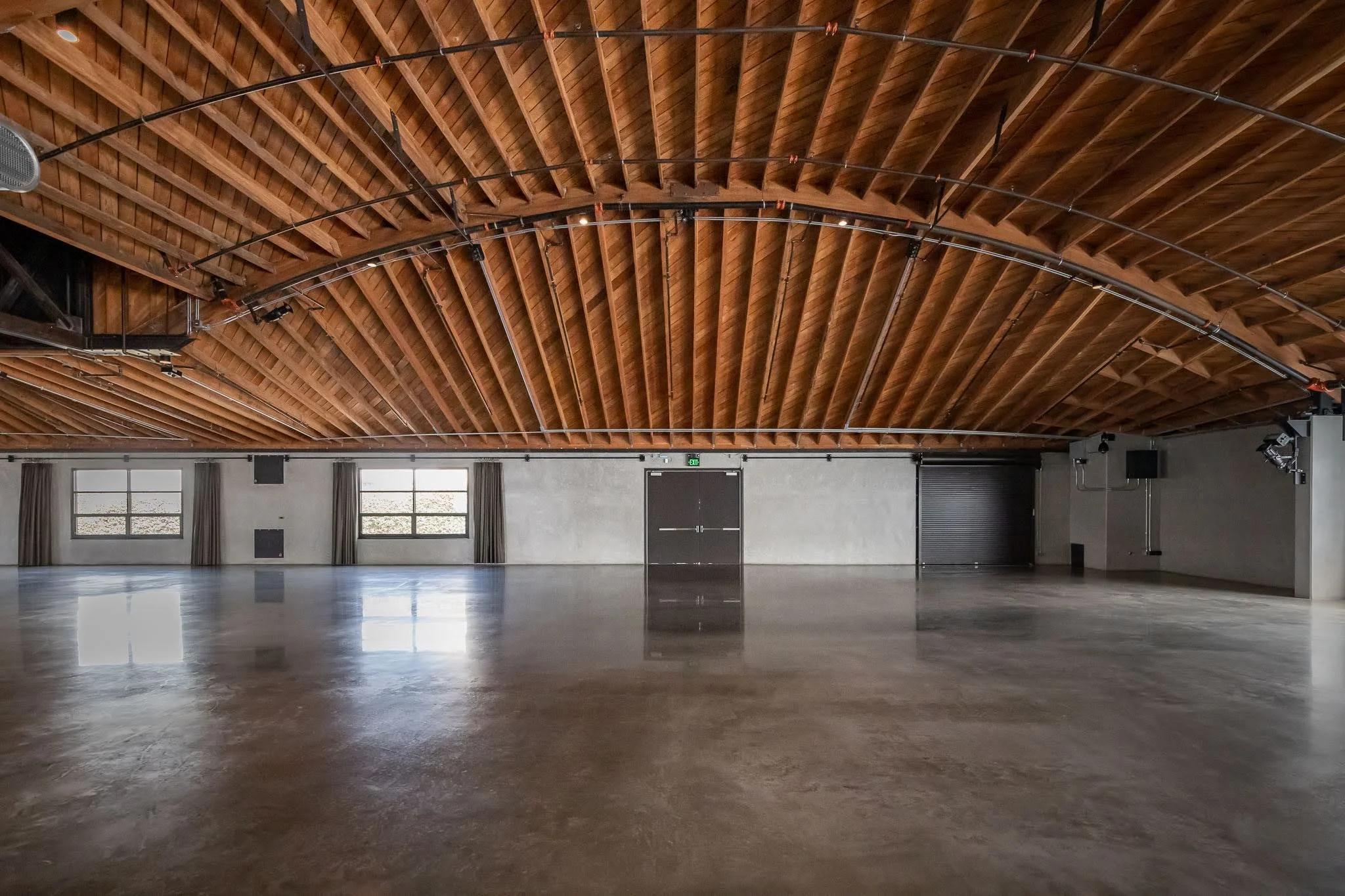 Empty indoor event or exhibition hall with wooden arched ceiling, a polished concrete floor, and three windows with curtains