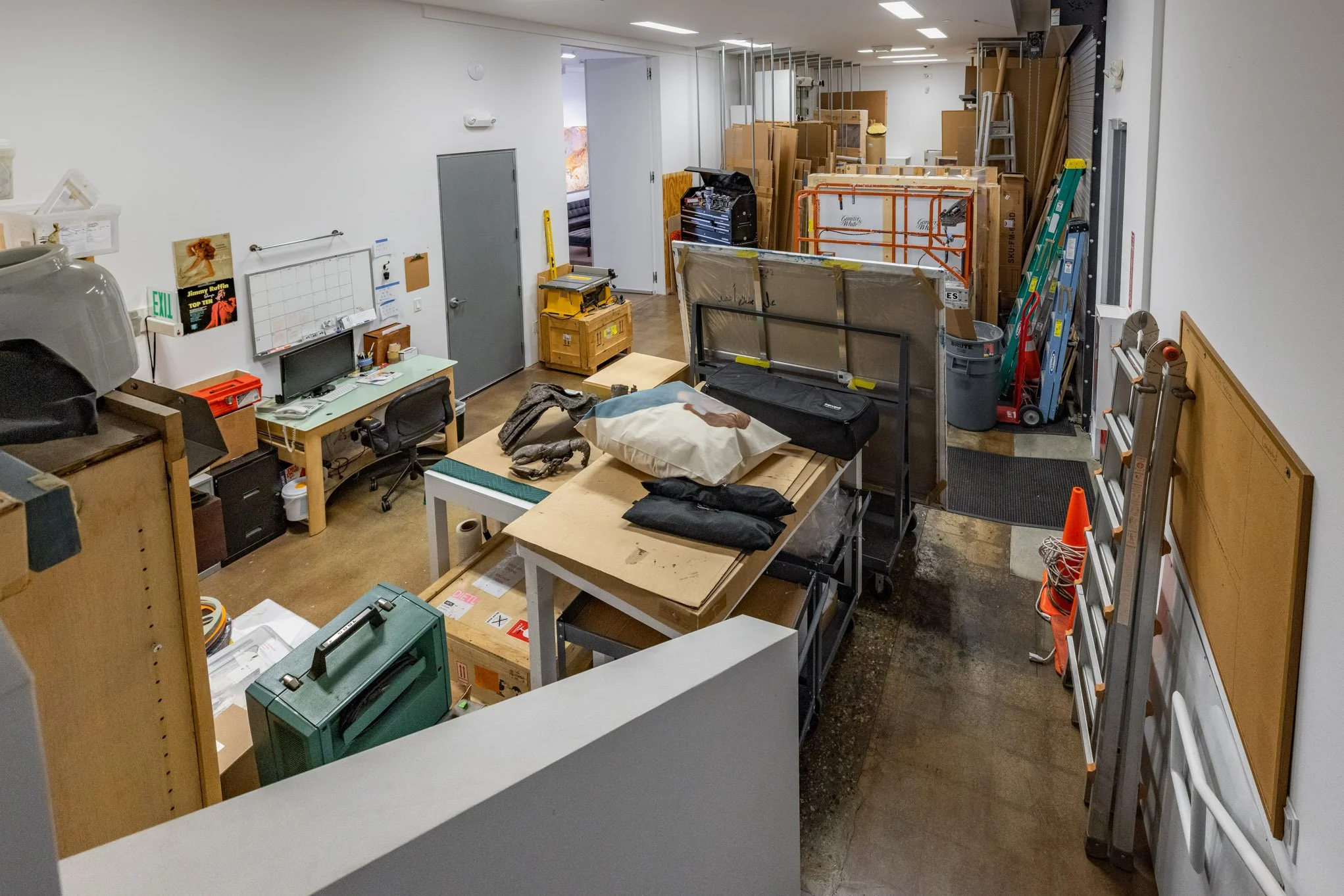 A cluttered workshop or storage room with work tables, tools, ladders, and storage boxes.