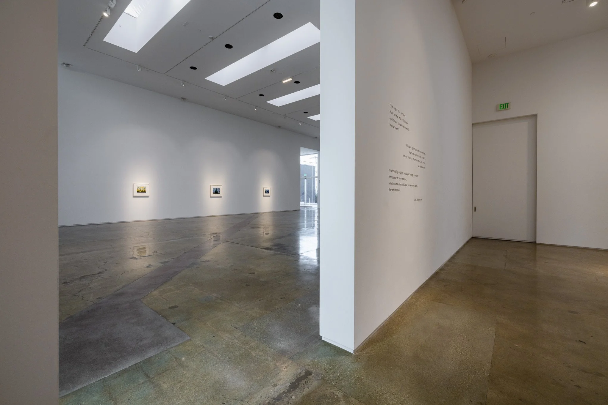 Interior of an art gallery with white walls, wooden and concrete floors, and ceiling skylights. Three framed artworks hang on the back wall, and a partial wall with a poem or exhibit description is on the right side near an emergency exit door.