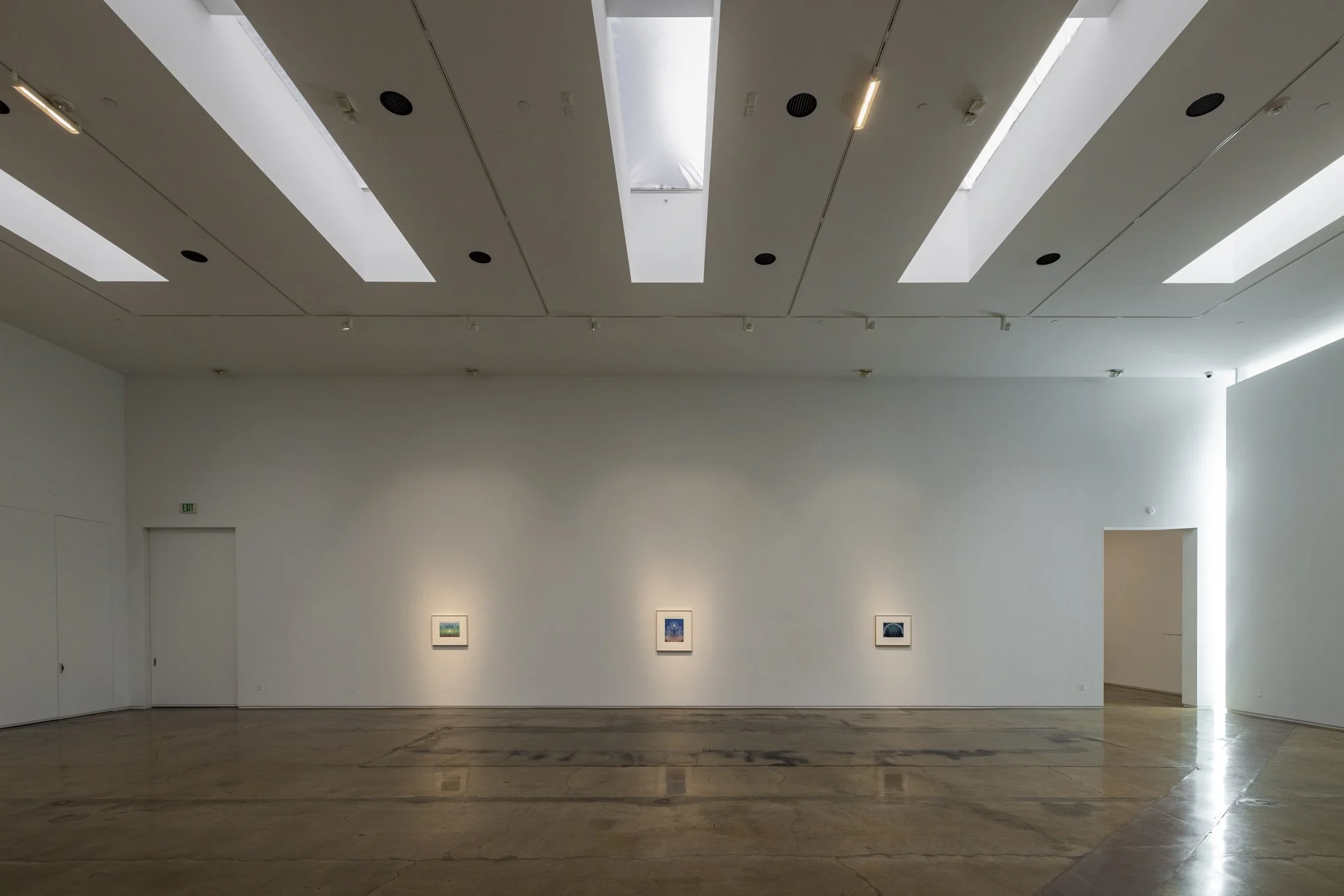 An art gallery with a white wall displaying three framed artworks illuminated by spotlights, with a minimalist ceiling design featuring skylights and a polished concrete floor.