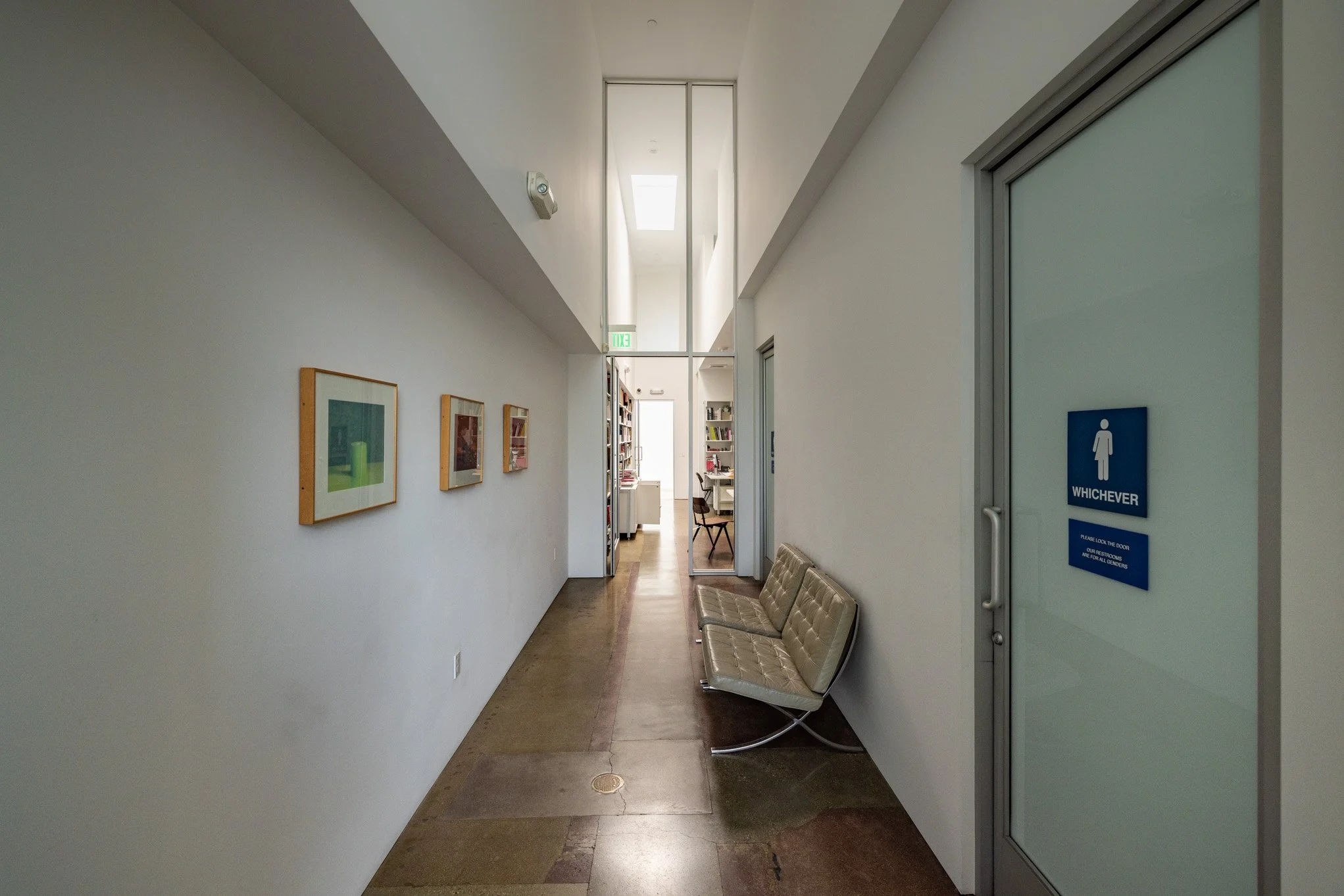 Interior hallway of a modern building with white walls, framed artwork, a beige bench, glass doors, and a bookshelf visible in the background.