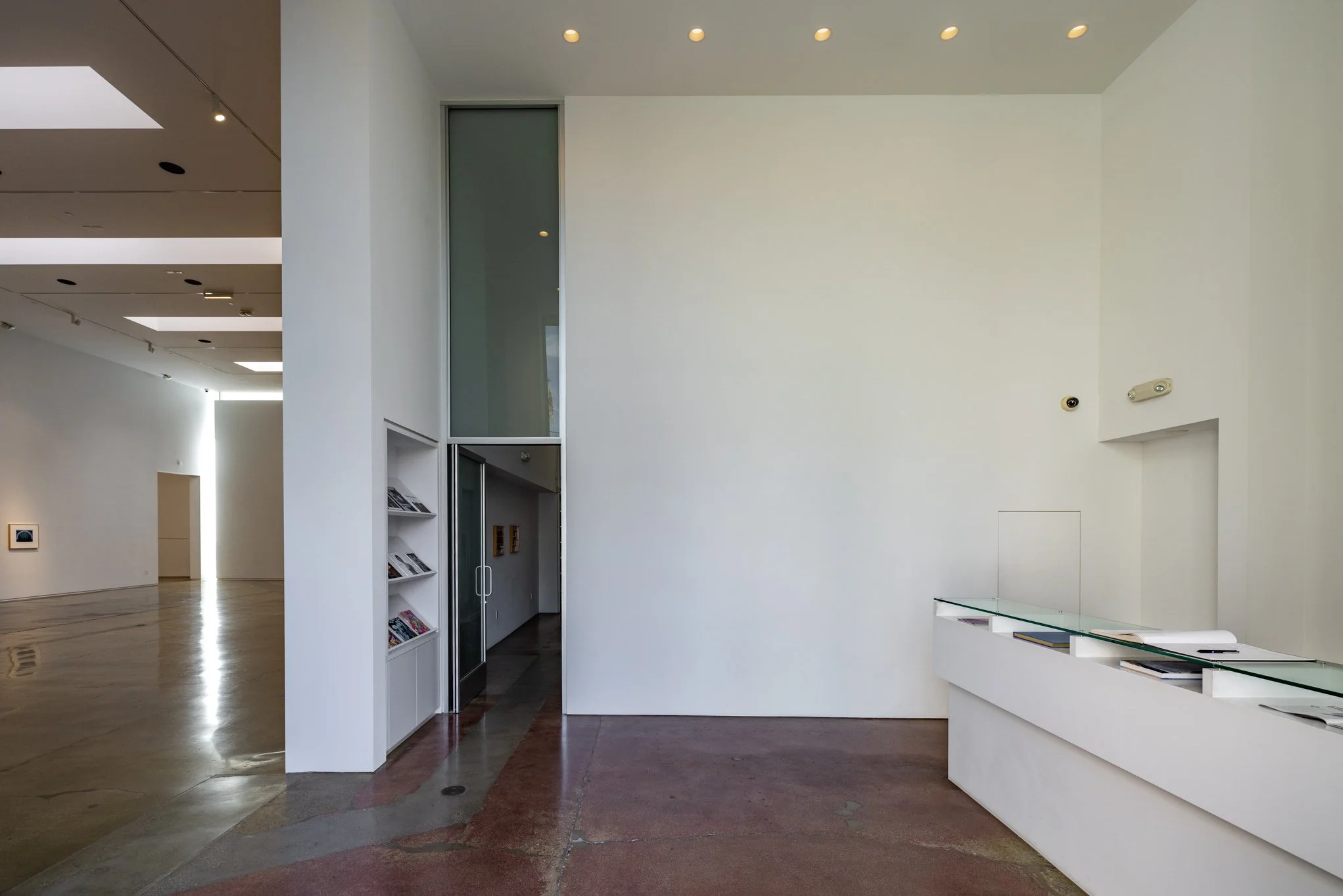 Modern art gallery interior with white walls, brown polished concrete floor, and a glass display case with books and papers. There are several small ceiling lights and an open doorway leading to another room with art on the walls.