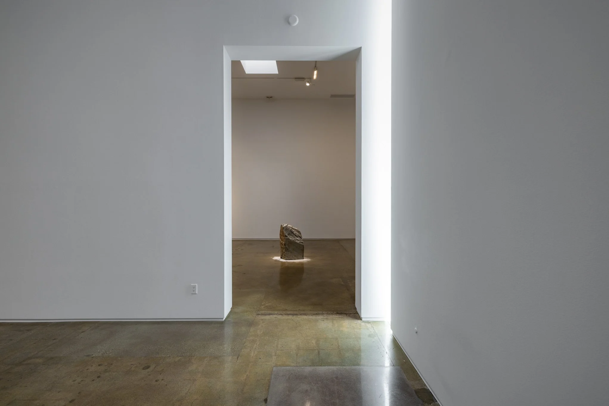 Empty art gallery with a single rock sculpture illuminated on the floor, viewed through a doorway with minimal lighting.