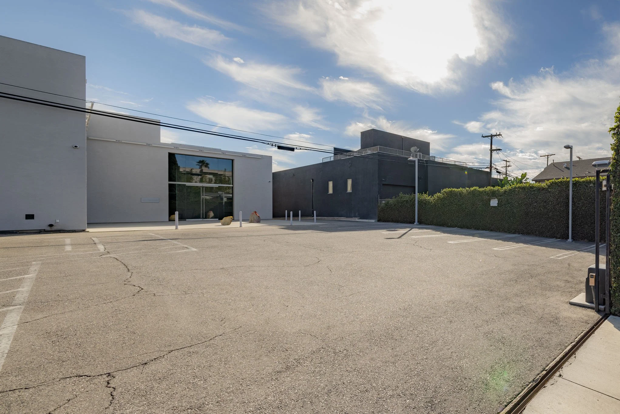Empty parking lot with cracks, modern white and black buildings, bushes, utility poles, and a partly cloudy sky.