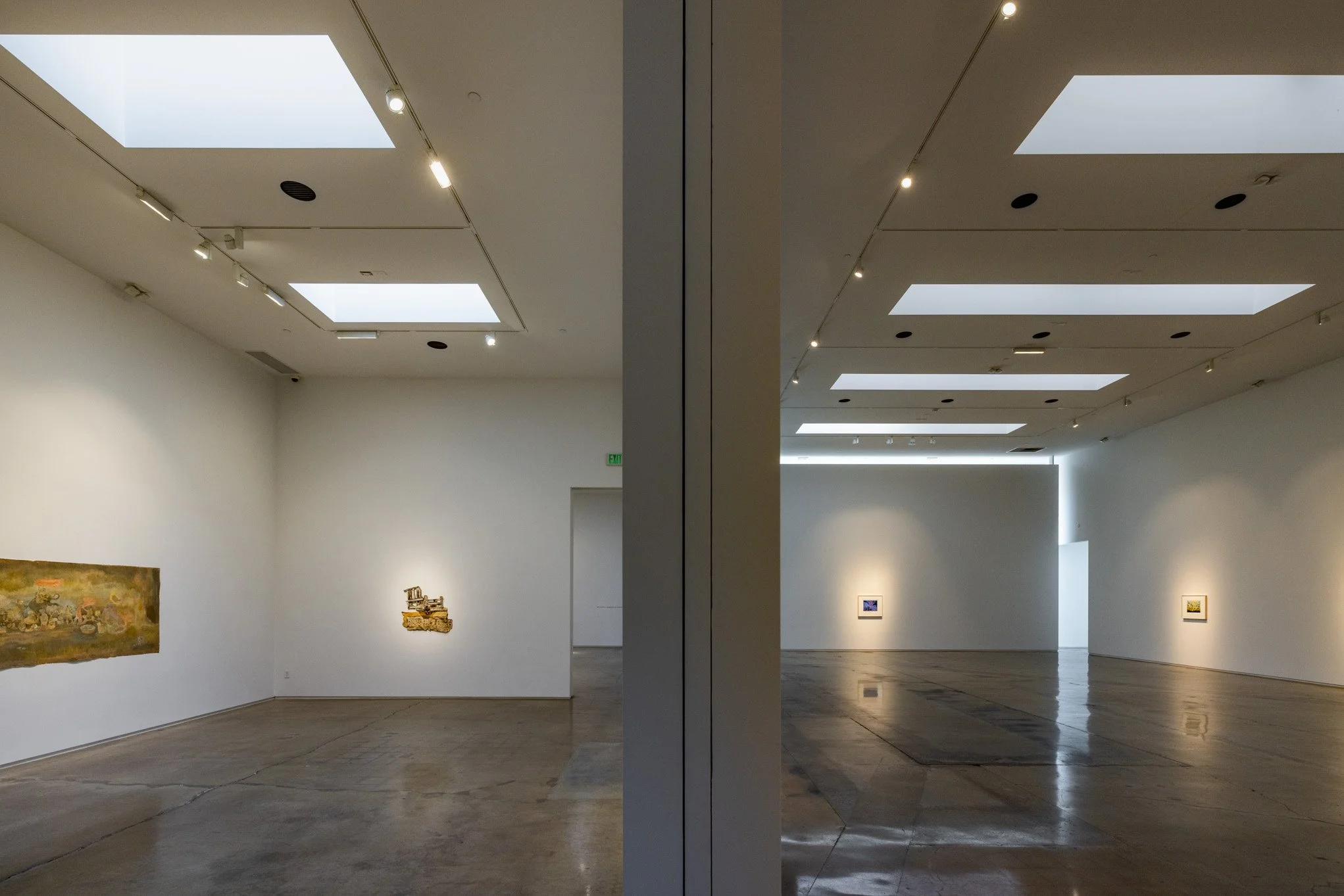 A diptych photo of an art gallery with white walls, hardwood floors, and skylights. The left side shows a large landscape painting, and the right side displays a small framed artwork on the wall. The middle vertical divider is a mirror reflecting the
