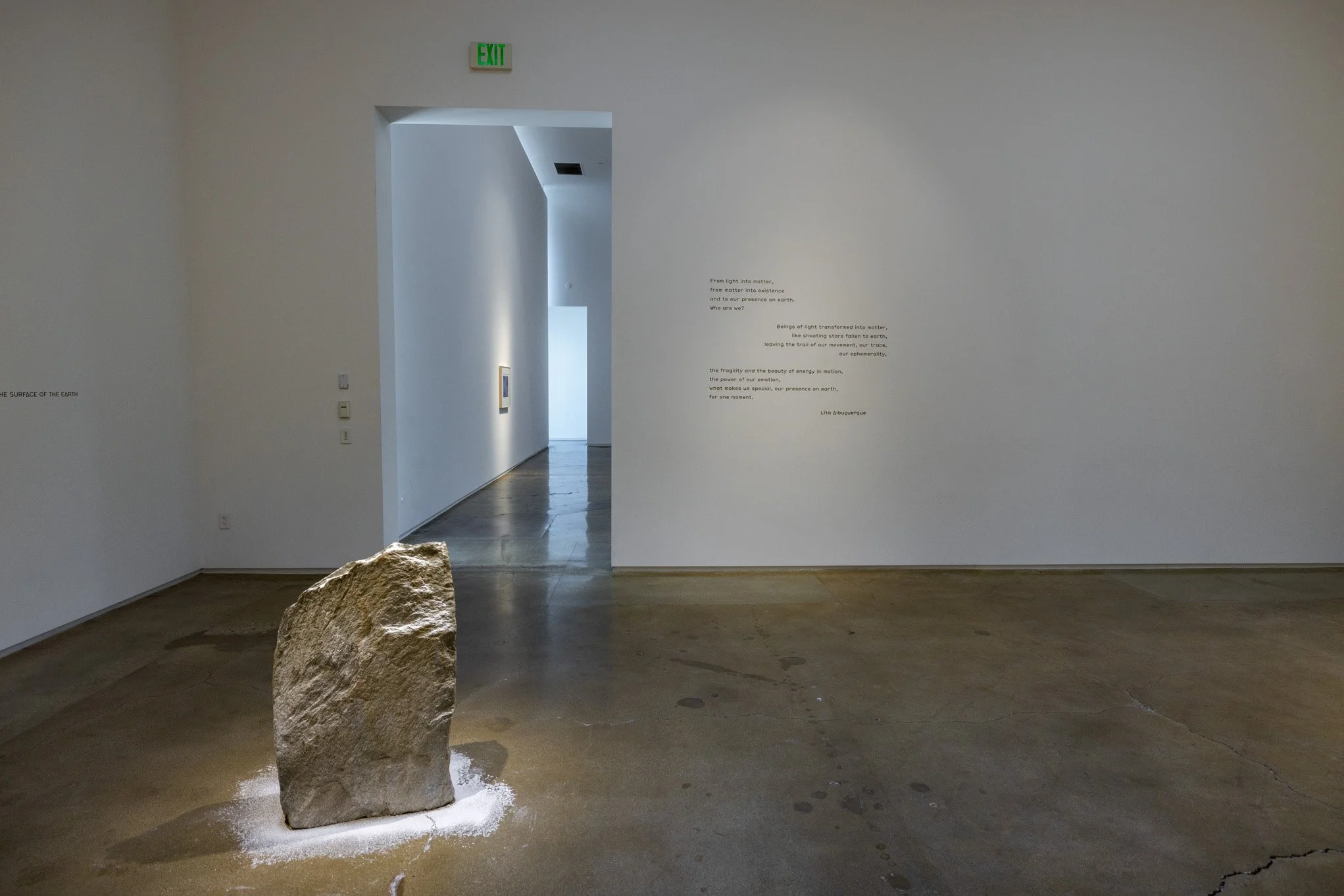 An art installation in a gallery featuring a large, rough stone with white powder underneath, placed on a polished concrete floor. The gallery walls are white with minimal text and artwork, and an exit sign is visible above a doorway.