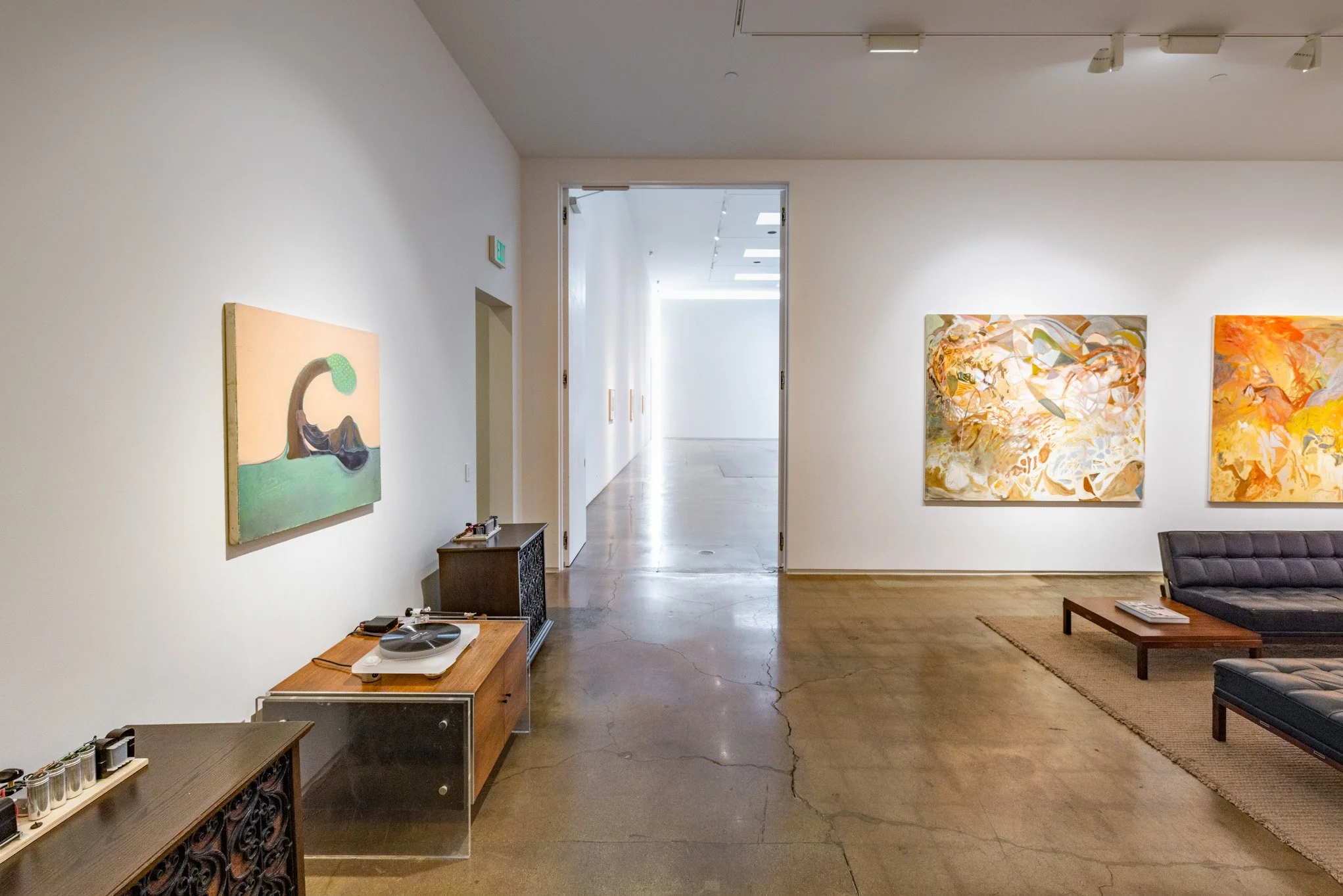 An art gallery with several abstract paintings on white walls, hardwood and concrete floors, a black sofa, a coffee table, and vintage-style furniture and equipment.
