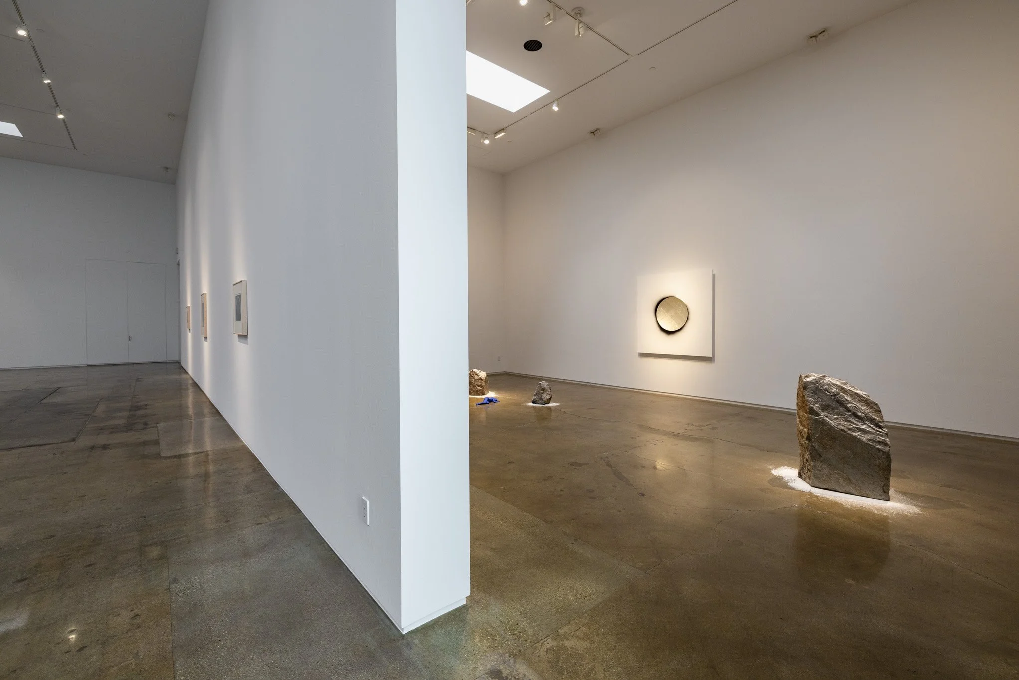A minimalist art gallery with white walls and polished concrete floors, displaying a circular artwork on the far wall and several large rocks placed on the floor in the foreground.