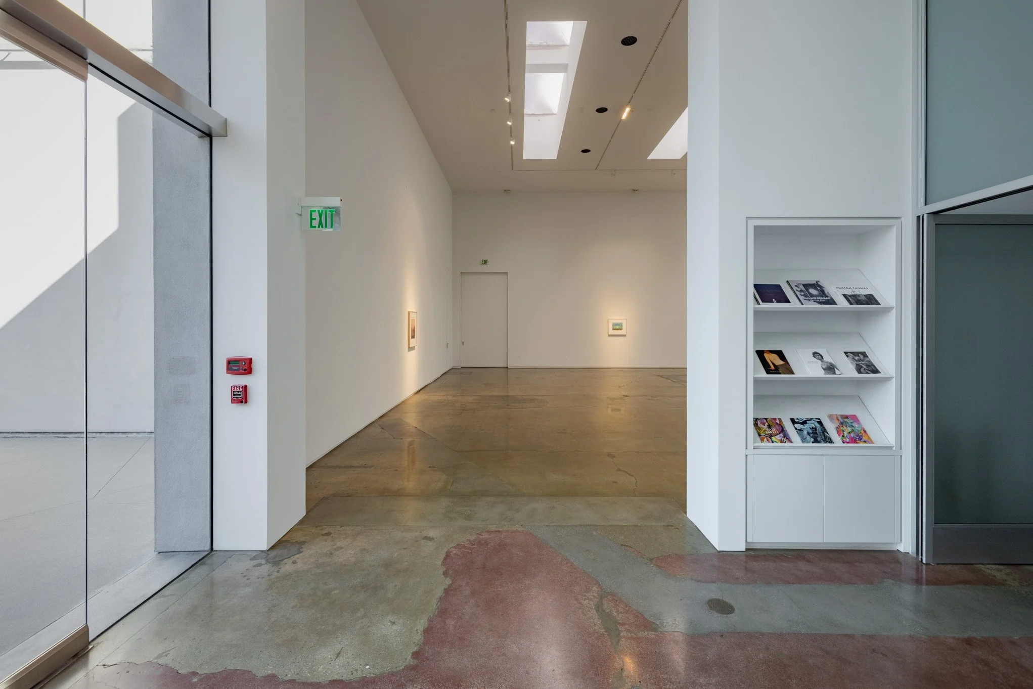 Empty art gallery with white walls, a door, framed artworks on the walls, a bookshelf with magazines, and a polished concrete floor.