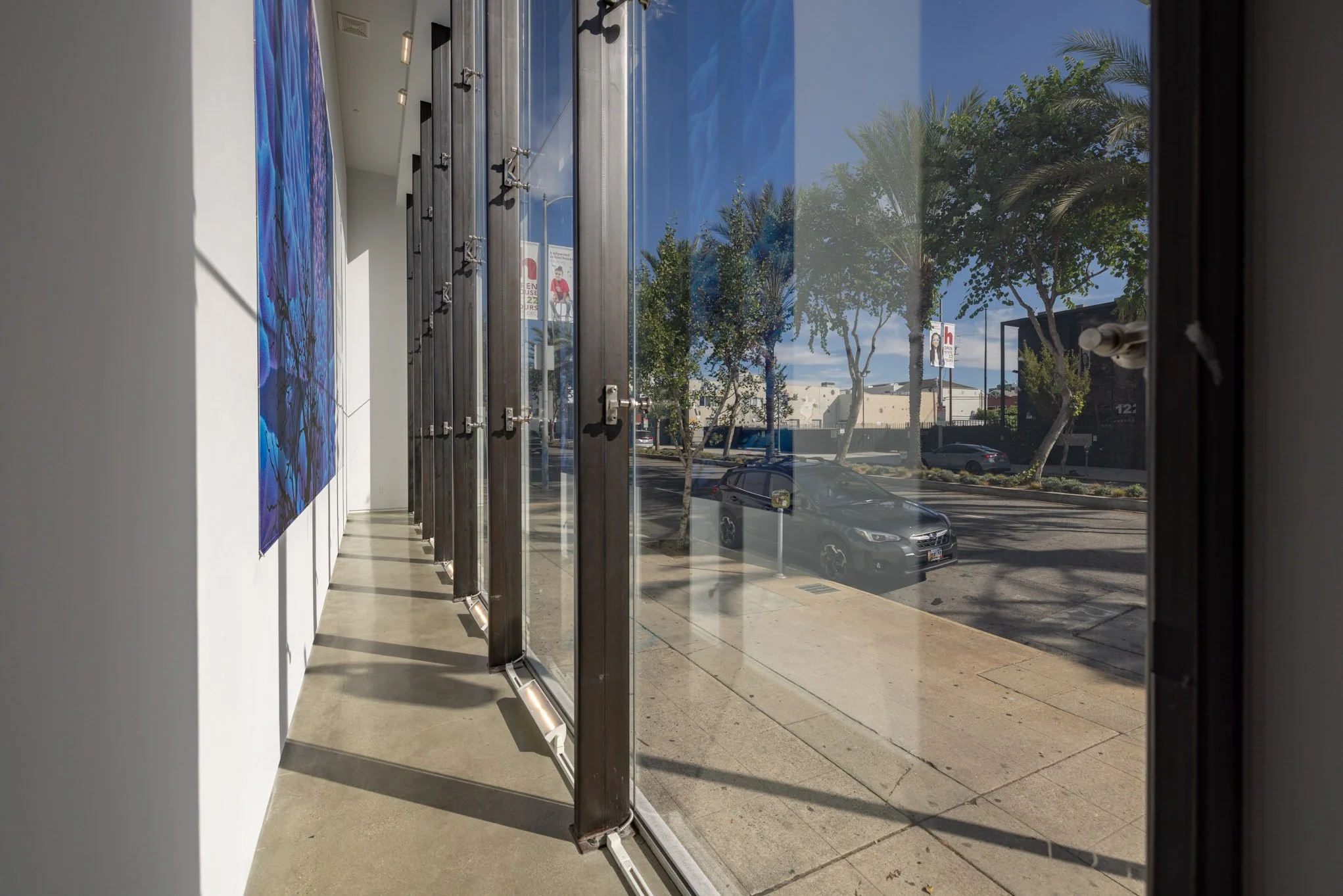 Interior view of an art gallery with a large blue abstract painting on the left wall, sunlight casting shadows on the polished concrete floor, and a large glass window showing trees, parked cars, and a street outside.