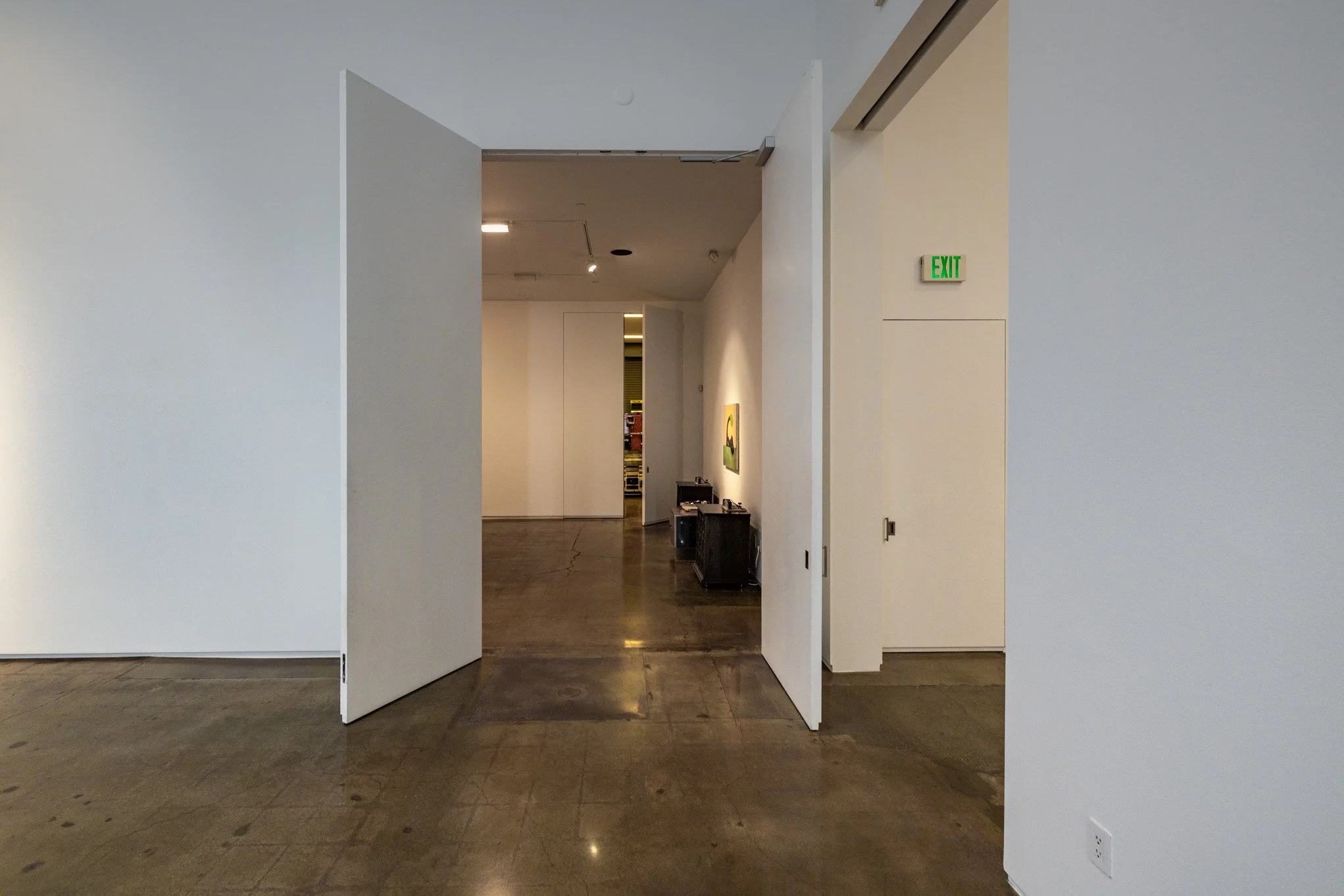 Empty art gallery with white walls, concrete floor, open white doors, and a small art piece on the wall inside.