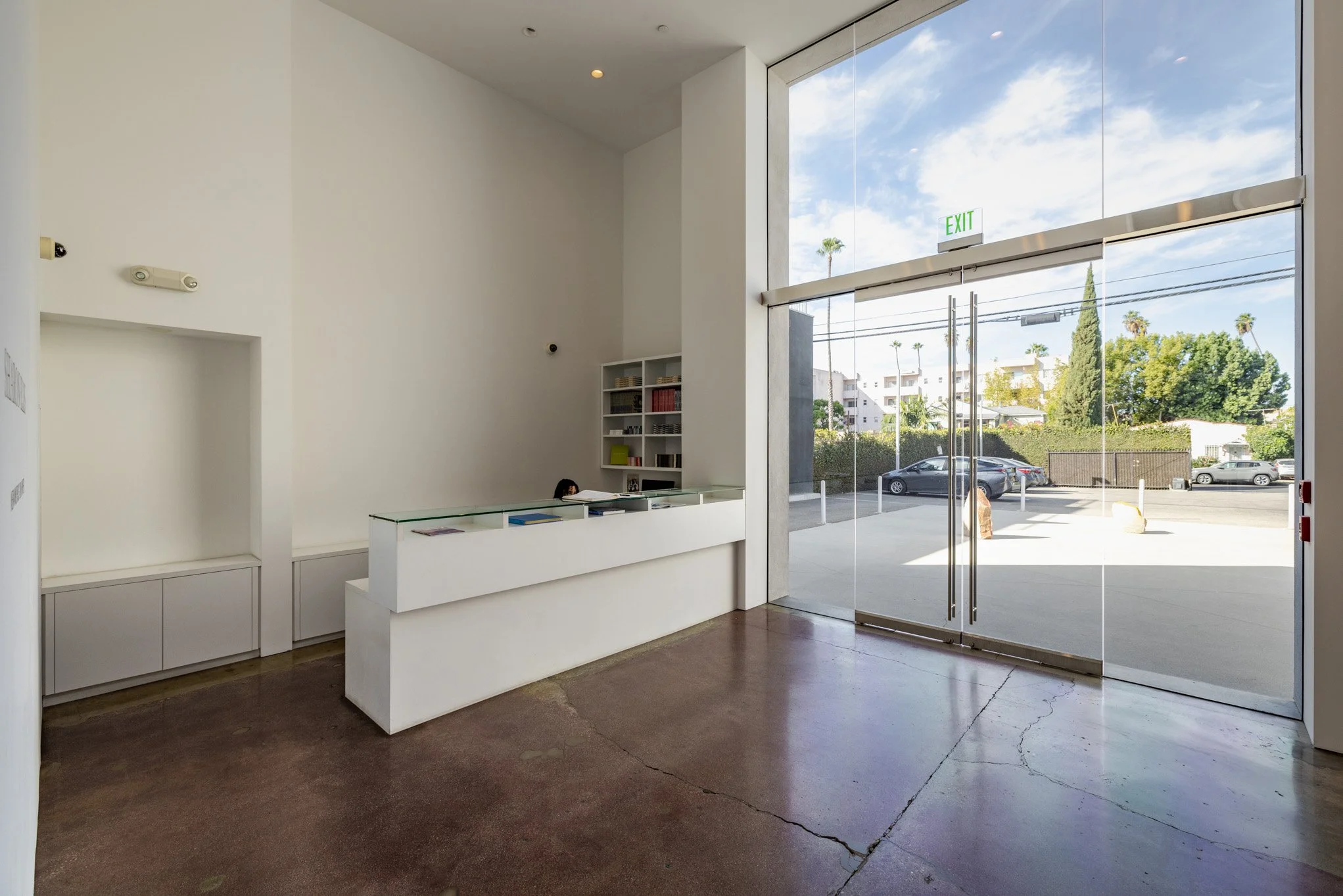 Empty modern reception area with a white desk, tall glass entrance doors, and large window showing a parking lot and trees outside.