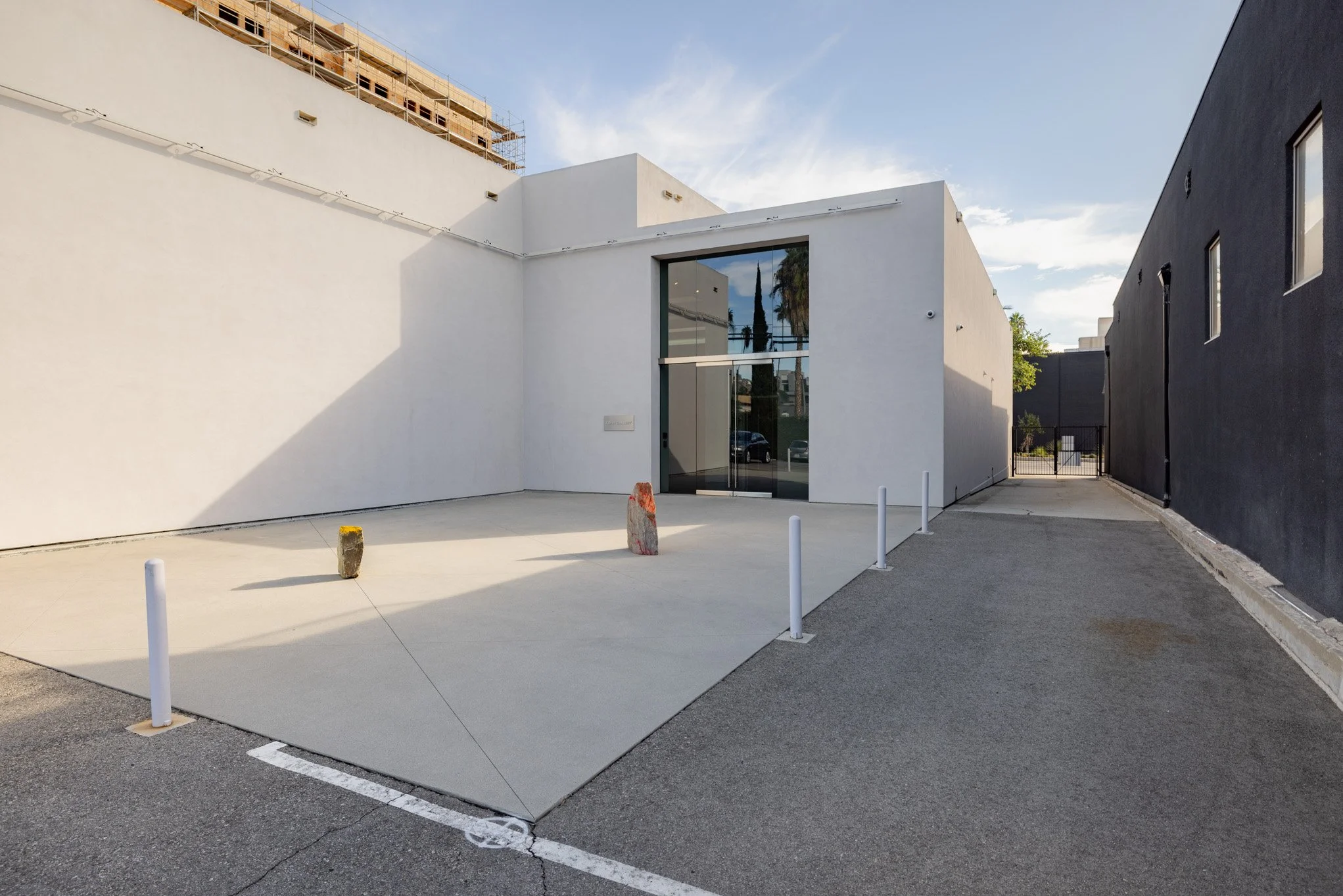 Modern white building with a large glass door, surrounded by parking lot and black walls, sunny day with clear sky.