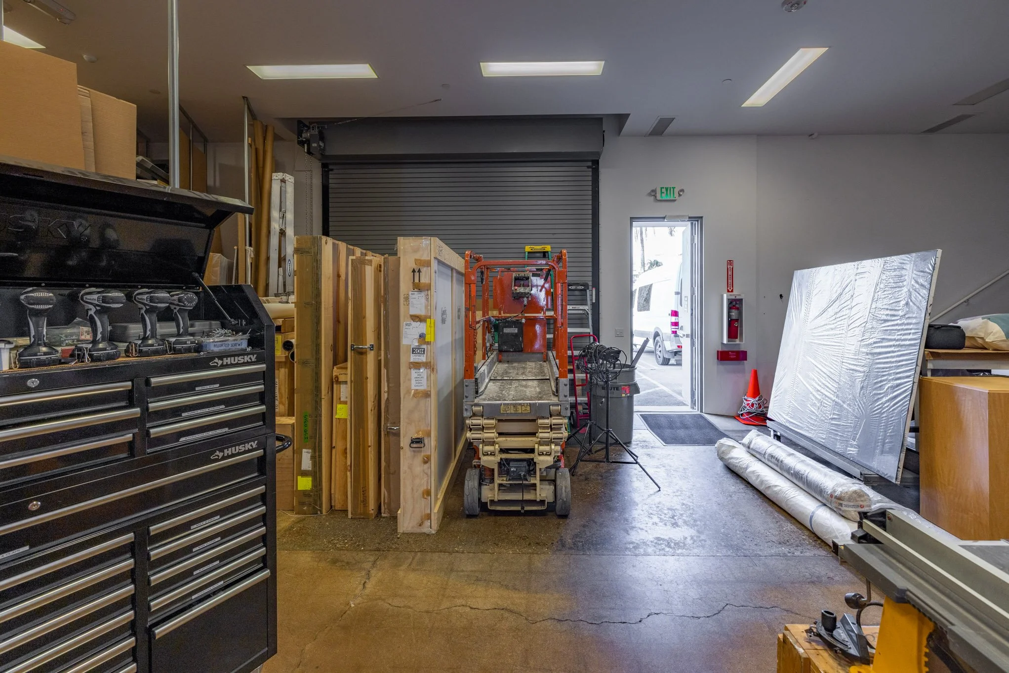 Workshop or storage area with tools, wood, and equipment, including a rolling scaffold, tool chest, and large metal sheets, near an open garage door and exit.