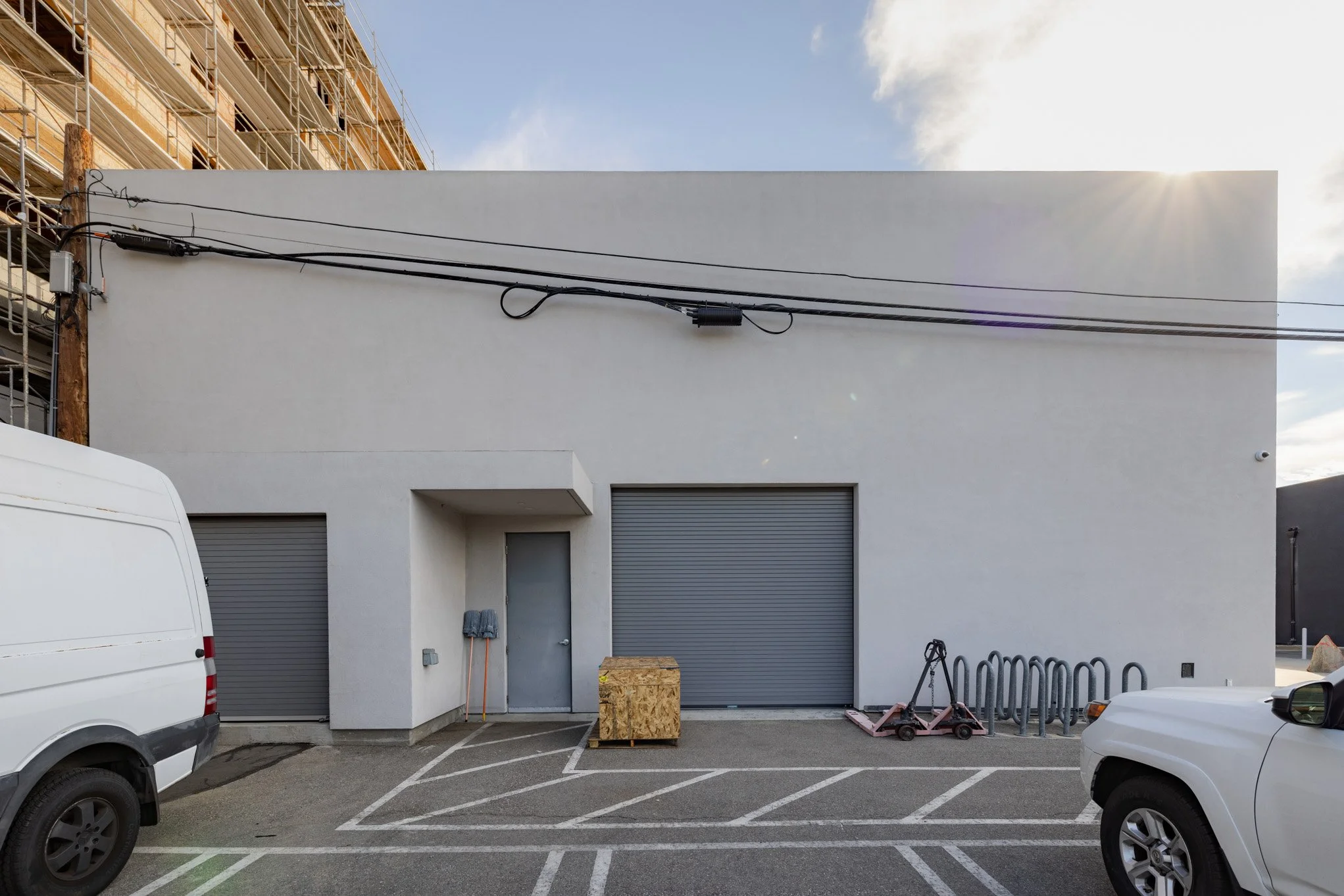 A plain white building with a small metal door and two large gray roll-up doors. A pallet jack and a wooden crate are in front. Two vehicles parked in front, and construction scaffolding on an adjacent building on the left. Power cables run across th