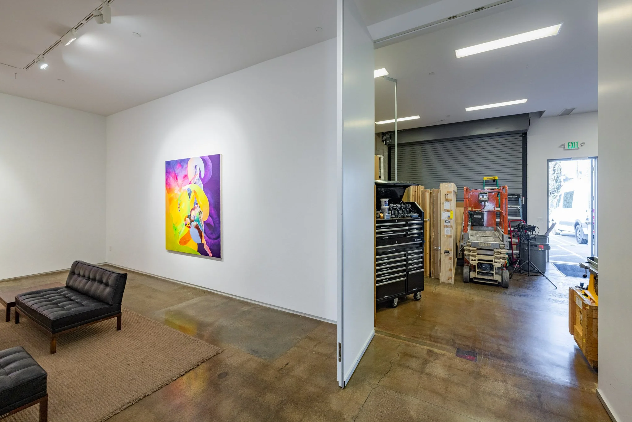 Interior view of an art gallery with a colorful abstract painting on the wall, black seating, and a partially open garage door revealing storage and construction equipment.