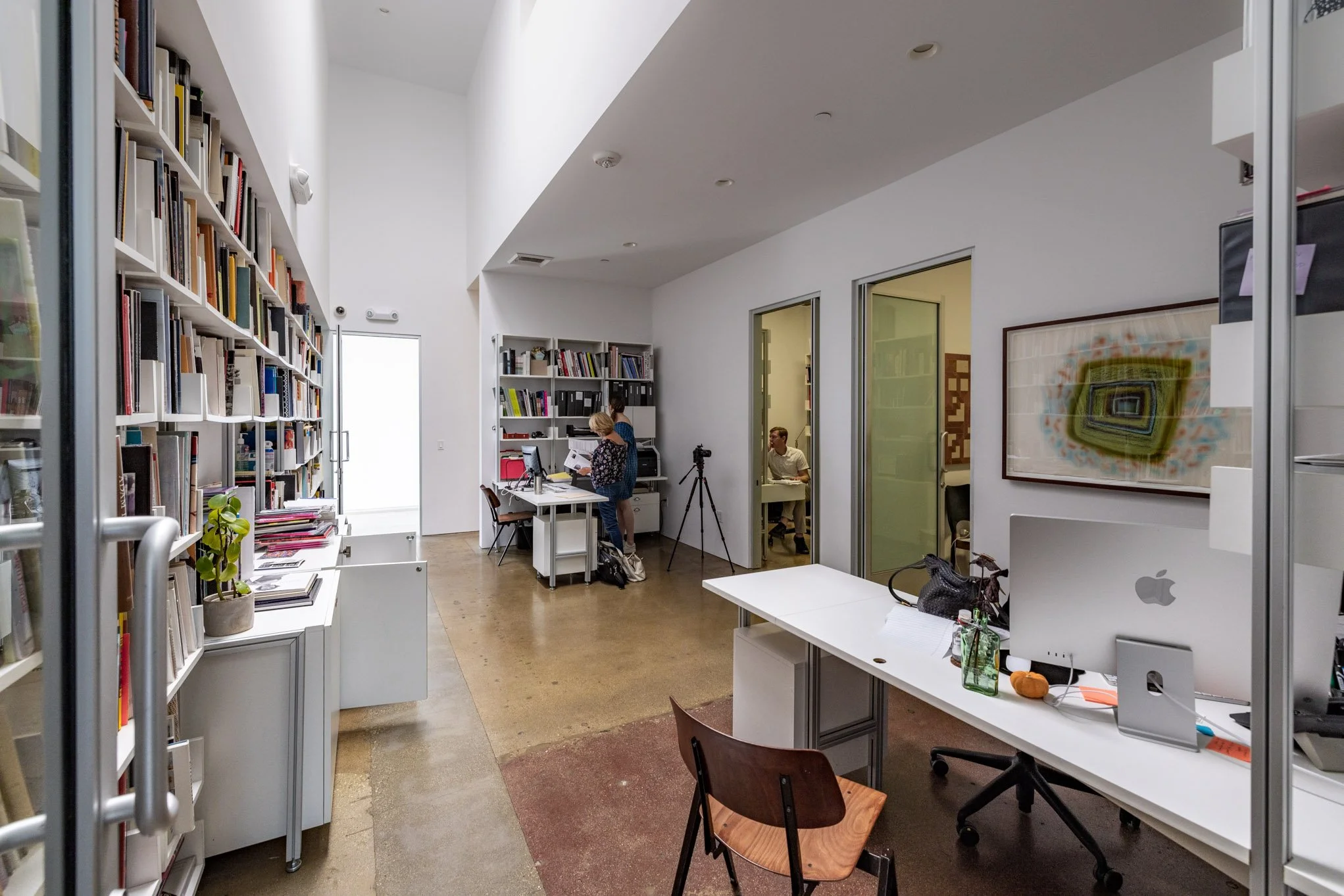 Interior view of a modern library or office with bookshelves filled with books, desks, chairs, and people working or studying.