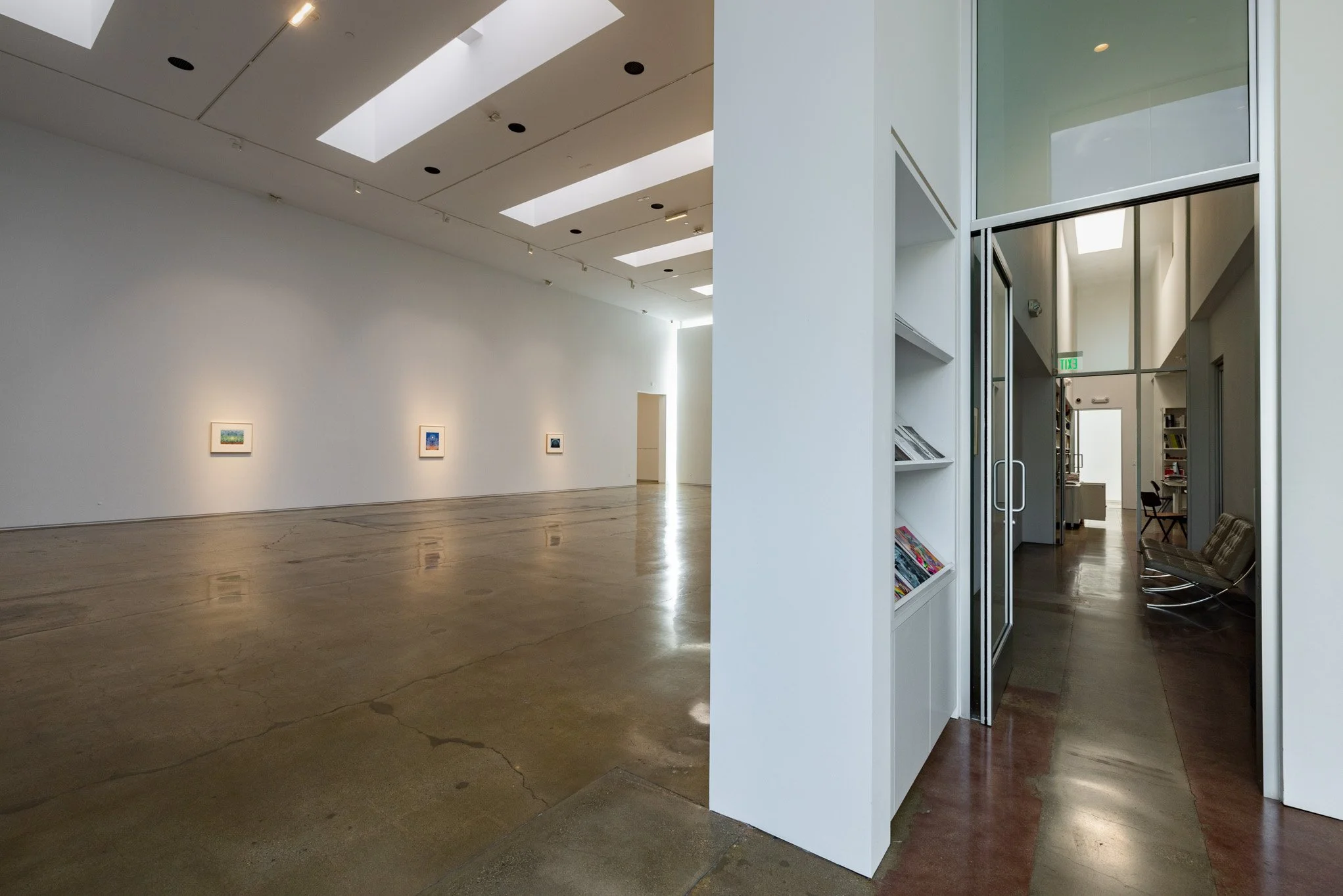 Wide empty art gallery with white walls, artwork on display, high ceiling with skylights, and polished concrete floor. Transition to a hallway with bookshelves and seating area.