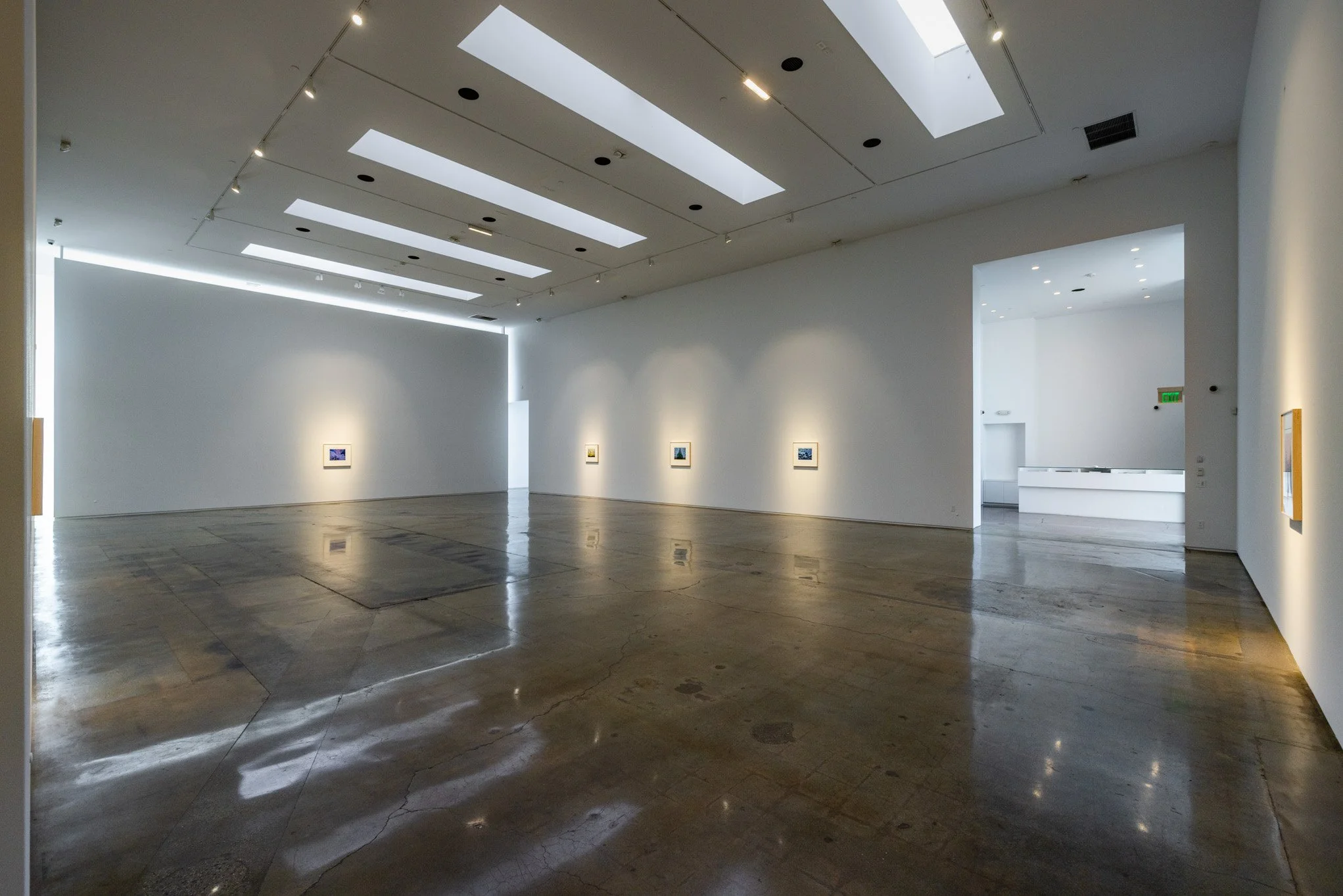 Empty art gallery with white walls and small framed artworks, polished concrete floor, bright ceiling lights, and natural light from skylights.