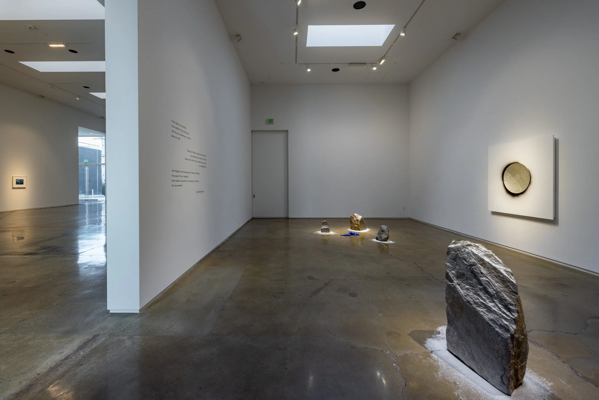 Contemporary art gallery with several rocks displayed on the floor, white walls, and abstract art on the walls. Natural light from skylights illuminates the space.