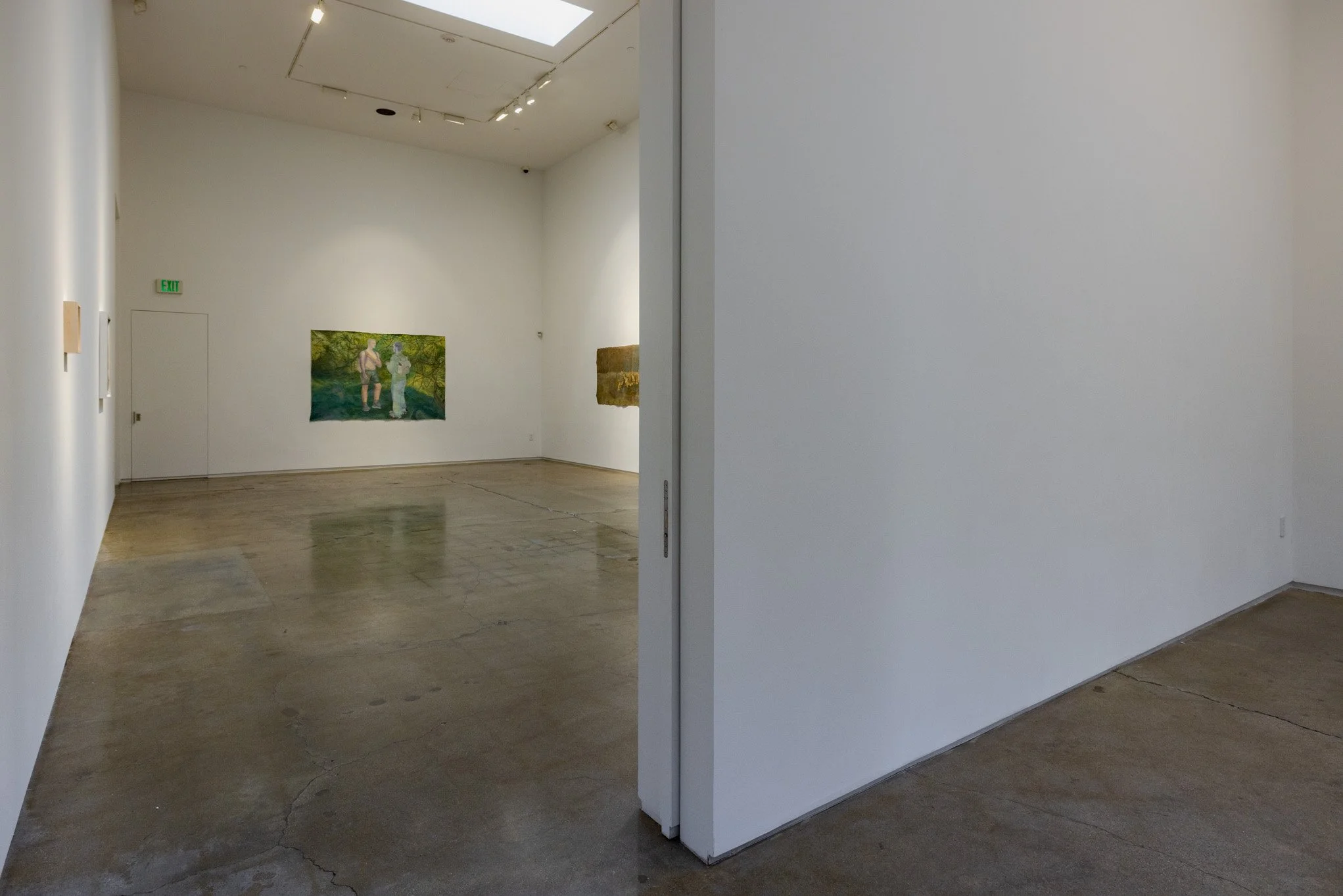 An art gallery with white walls, a concrete floor, and a doorway leading to another room. Some paintings are visible on the walls.