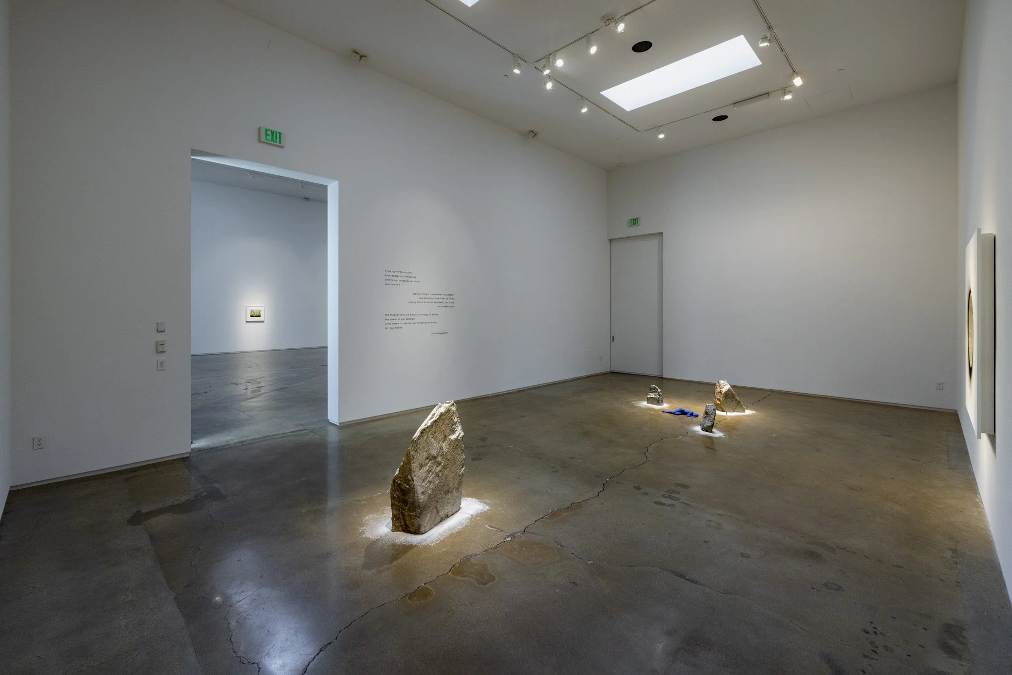 Art gallery room with three stone sculptures on the floor, lit by overhead lighting, with artwork hanging on the walls and text providing context.