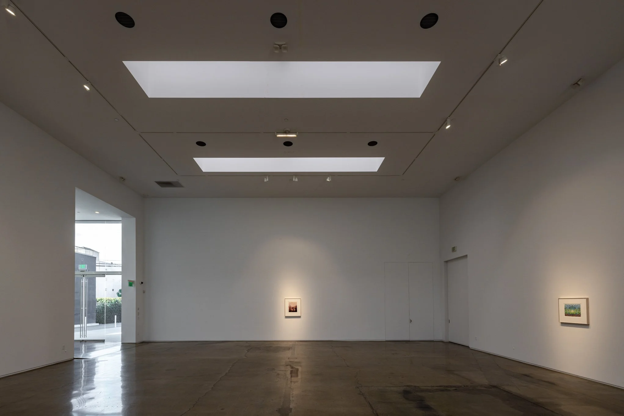 Empty art gallery with three framed paintings on white walls, polished concrete floor, and skylights in the ceiling.