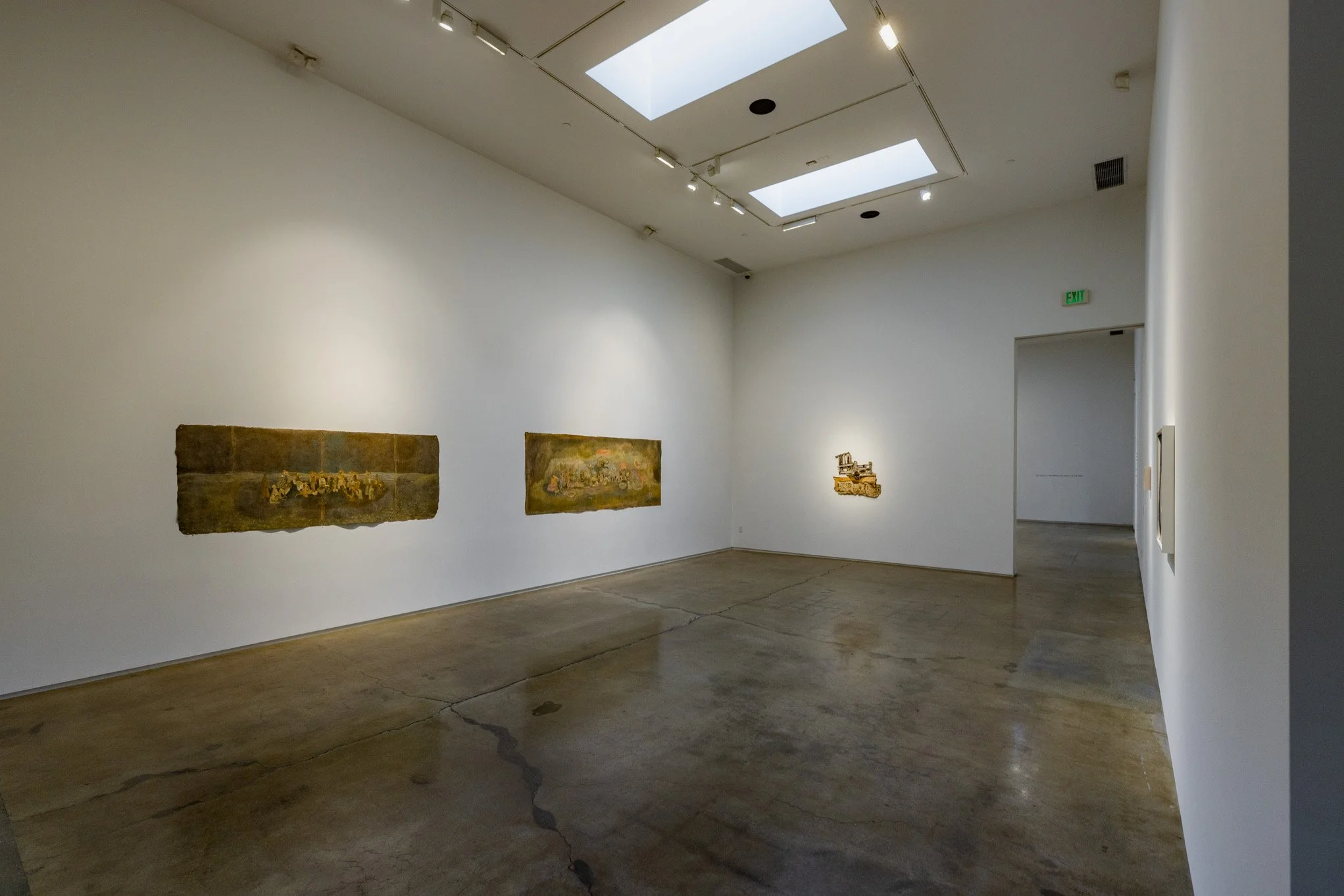 An art gallery with four paintings on white walls. The ceiling has skylights and track lighting, with a polished concrete floor.
