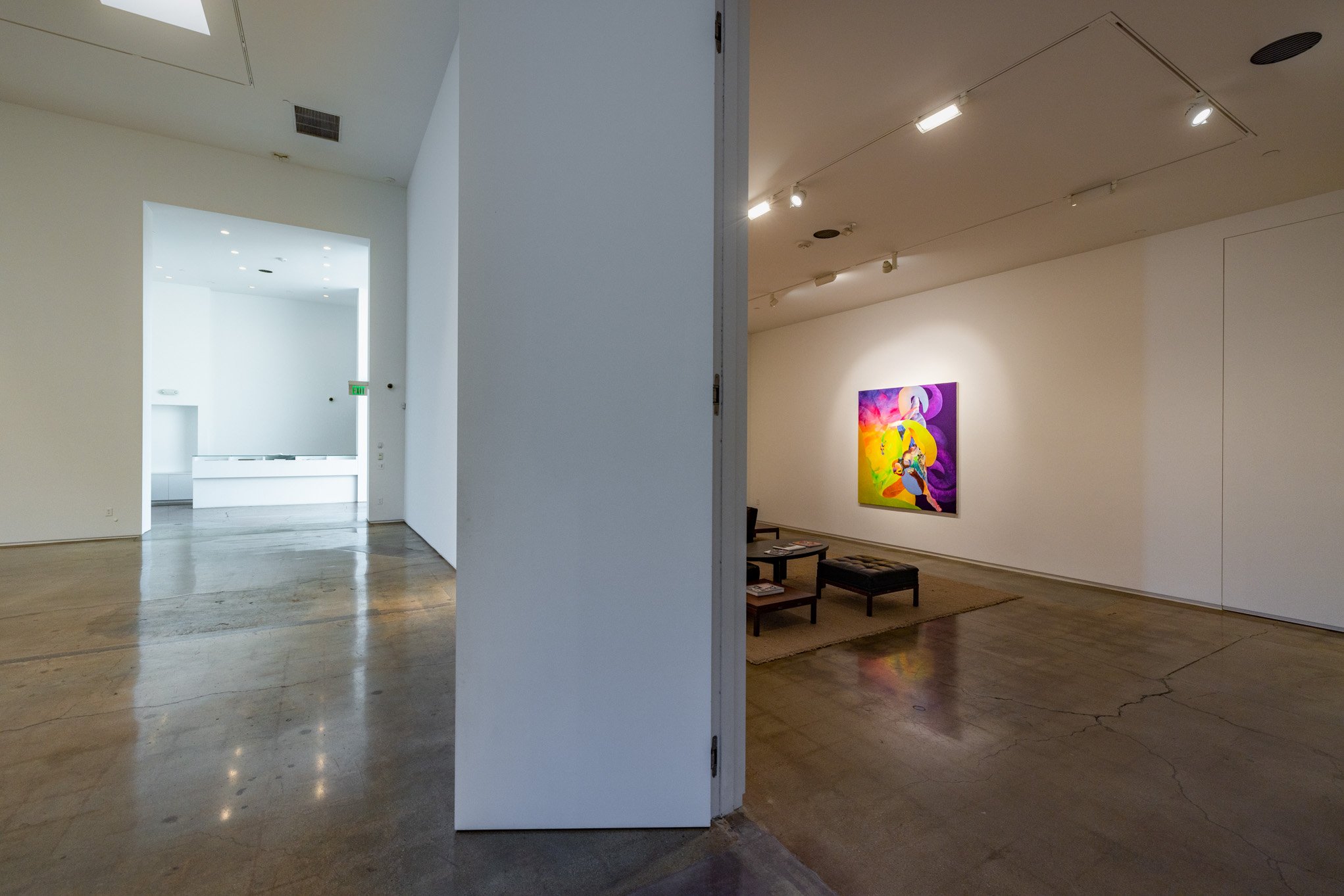Scene of two gallery rooms separated by a wall, with the left room being brightly lit and mostly empty, and the right room displaying a colorful abstract painting on a white wall, with seating and a rug below the artwork.