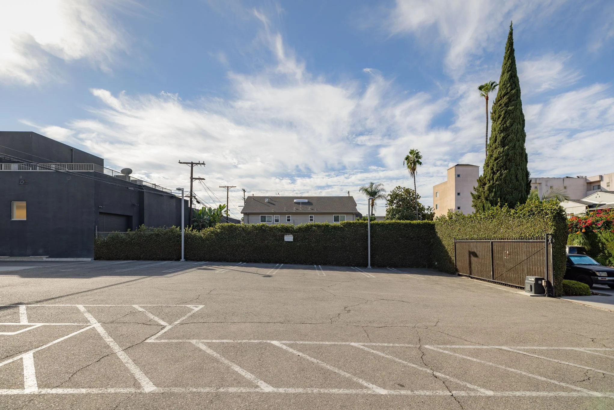 Empty parking lot with white painted lines, surrounded by hedges and buildings, under partly cloudy sky.