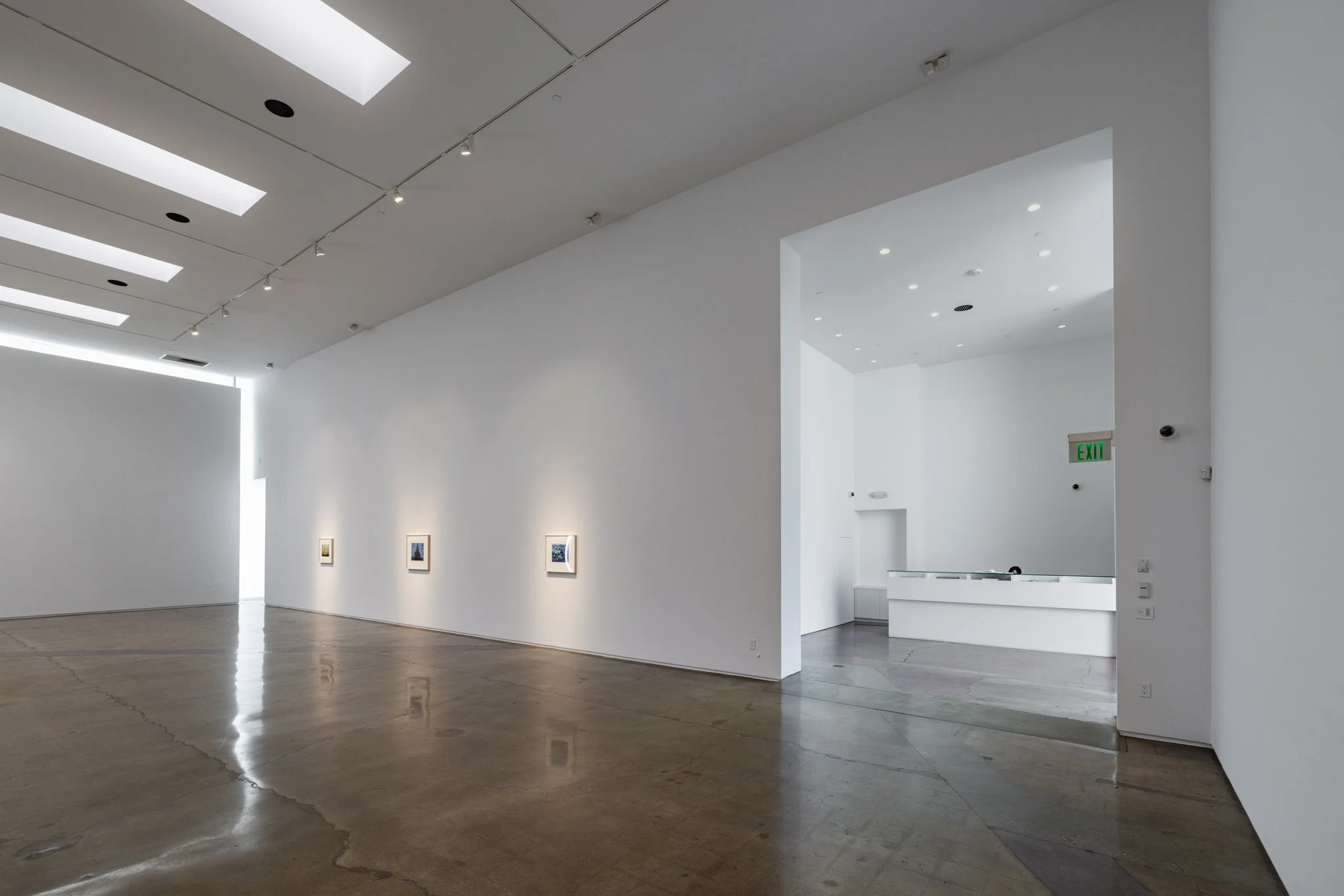 Interior view of an art gallery with white walls, polished floors, a few small framed artworks on the left wall, and an illuminated exit sign above a doorway on the right.