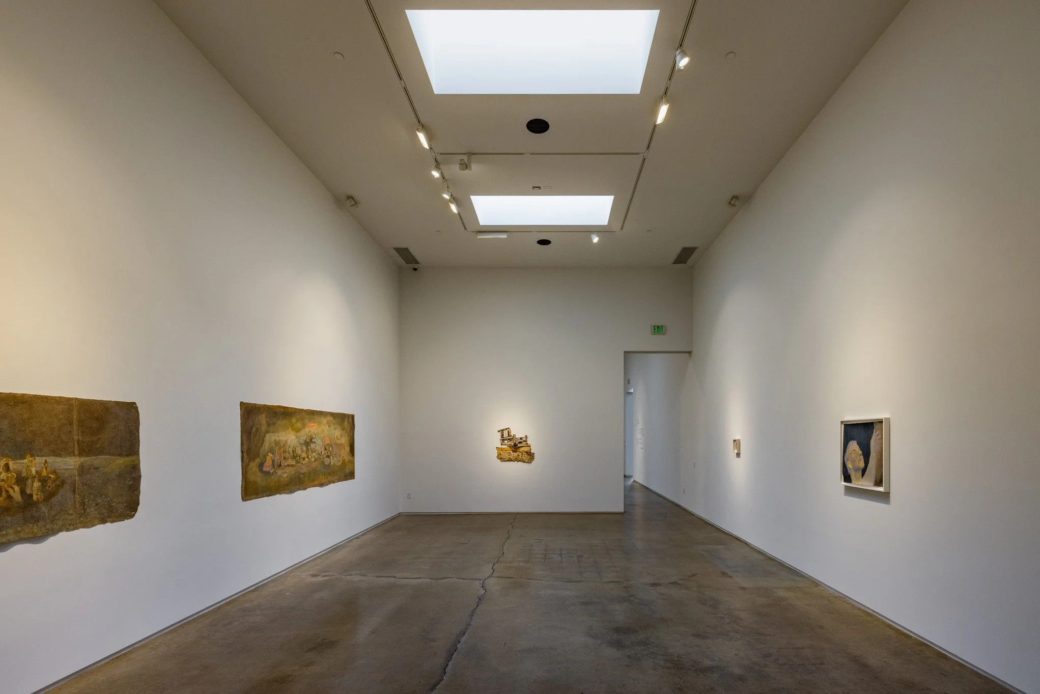 Empty art gallery with white walls, concrete floor, and paintings hung on the walls. Two large skylights in the ceiling provide natural lighting.