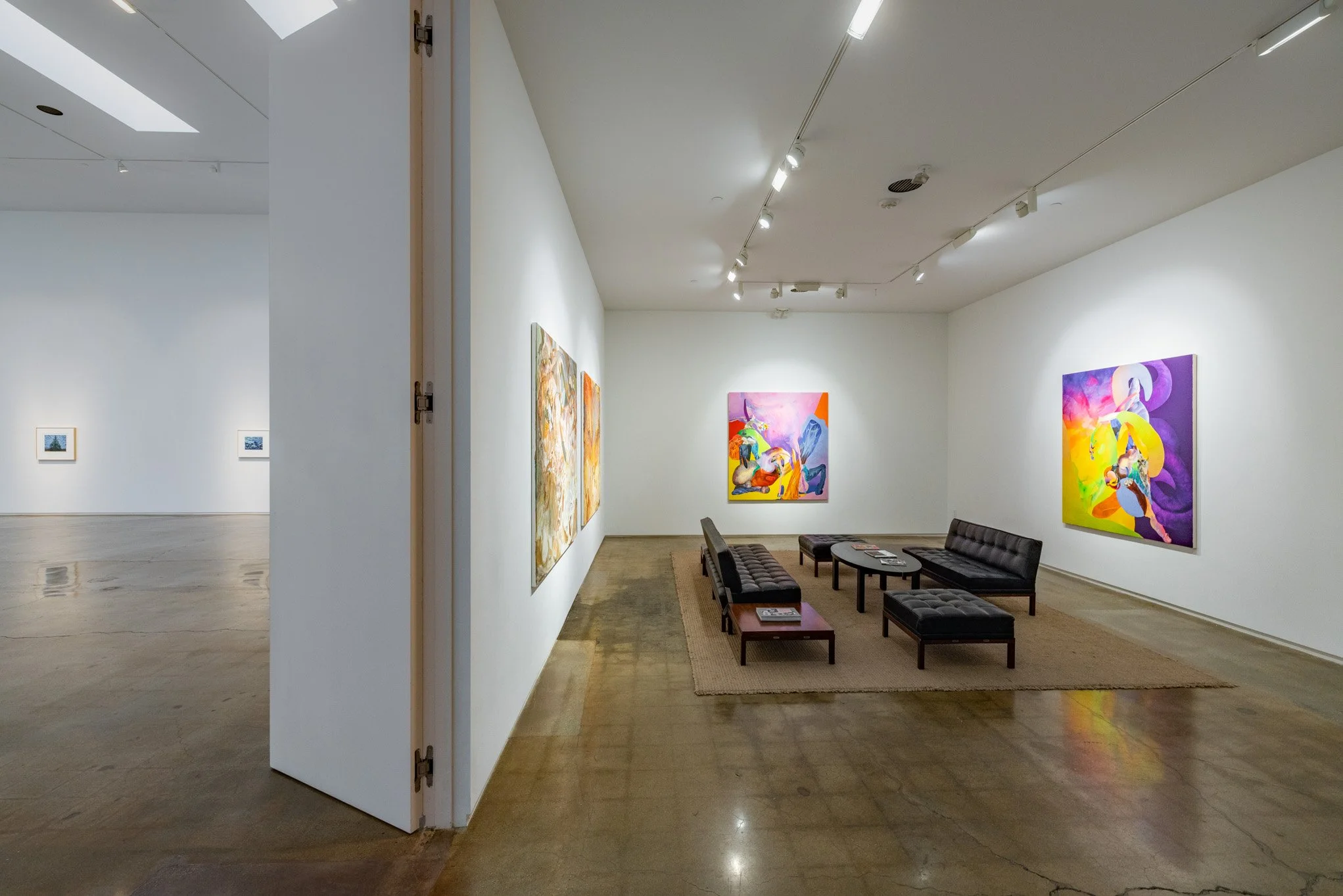 Interior of an art gallery with white walls displaying colorful paintings, a seating area with black leather sofas, and wooden coffee tables on a beige rug.