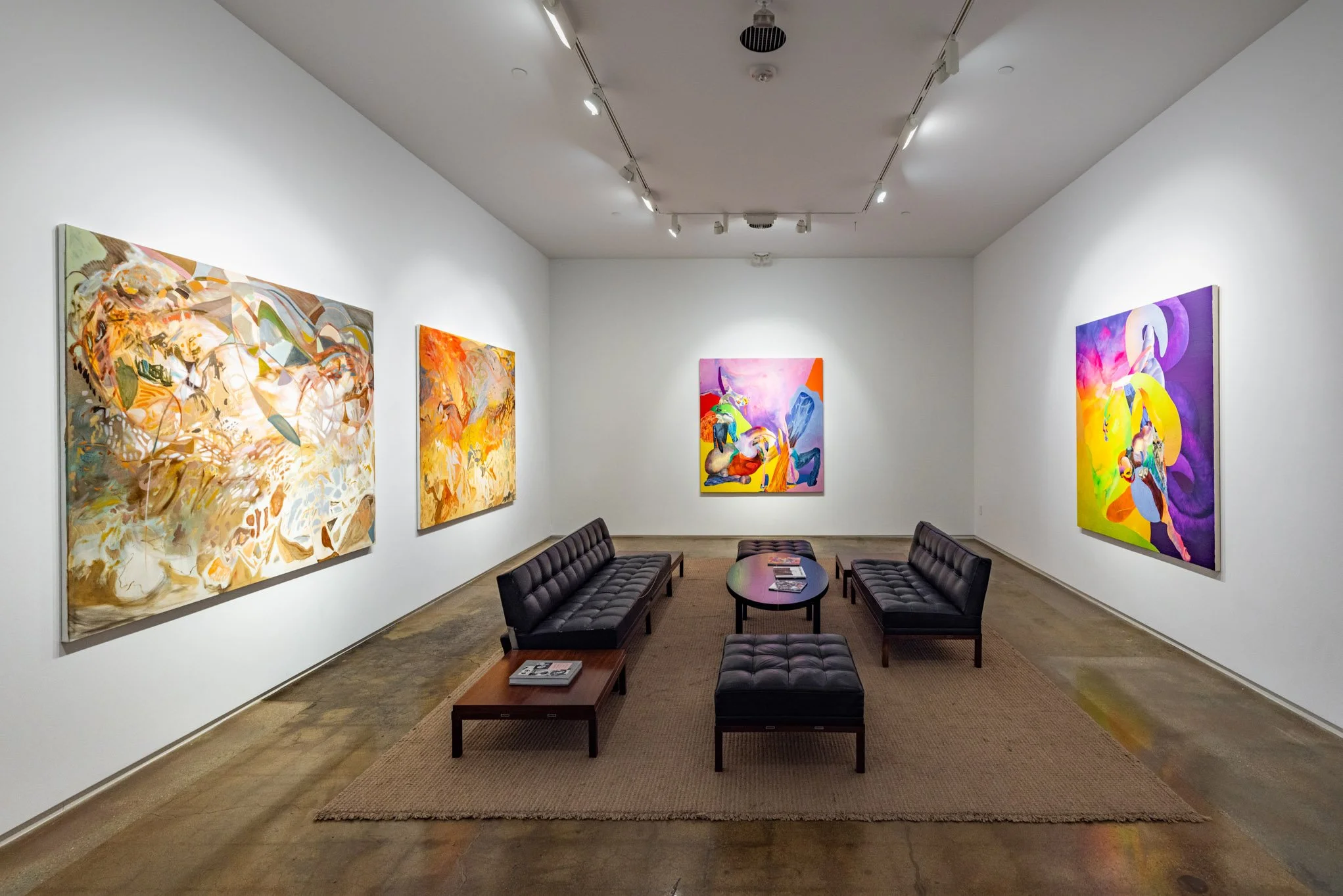 An art gallery featuring four colorful abstract paintings on white walls, with black leather couches and a small table in the center, on a brown rug.