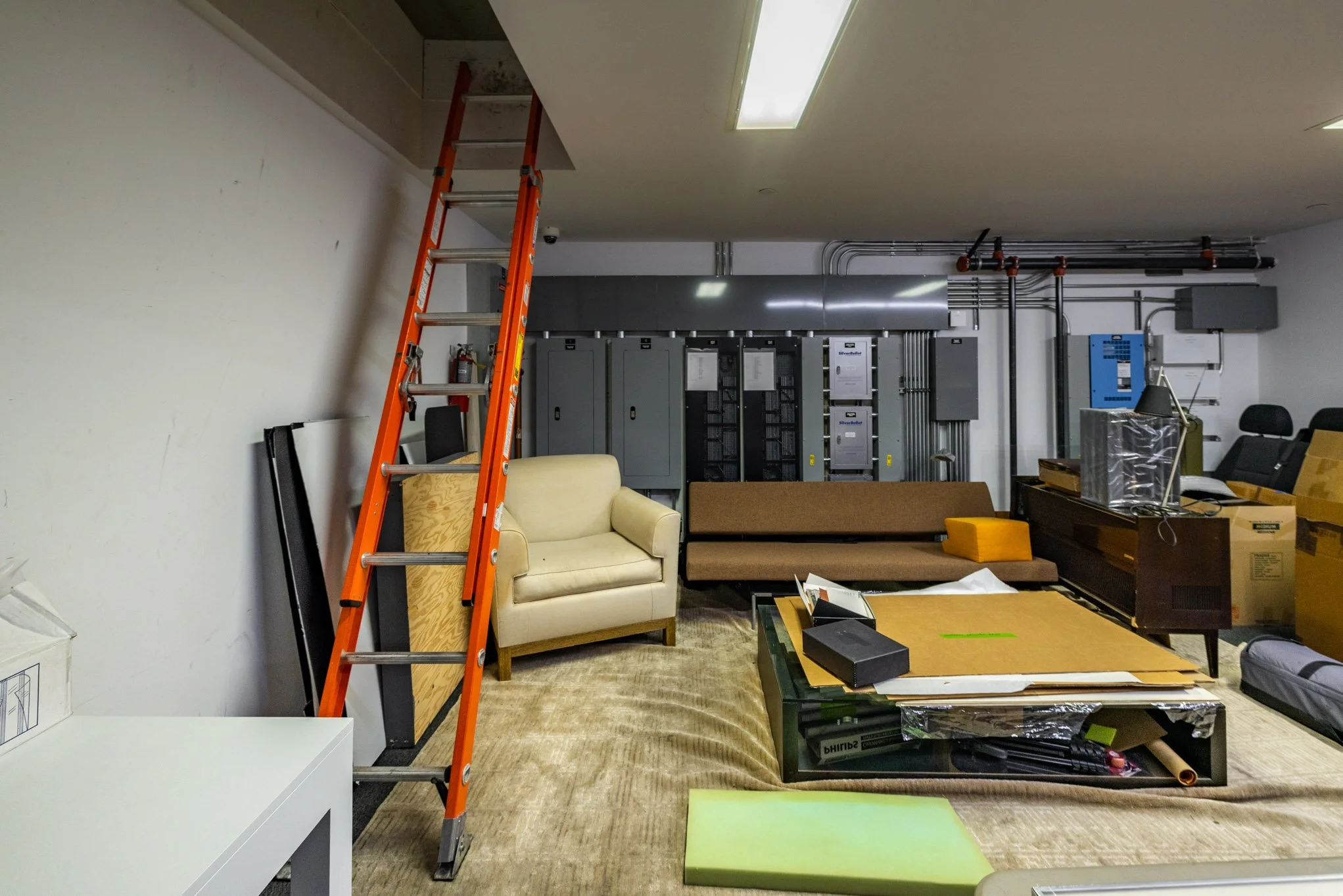 A cluttered room with furniture, boxes, a ladder, and electrical panels on the back wall.