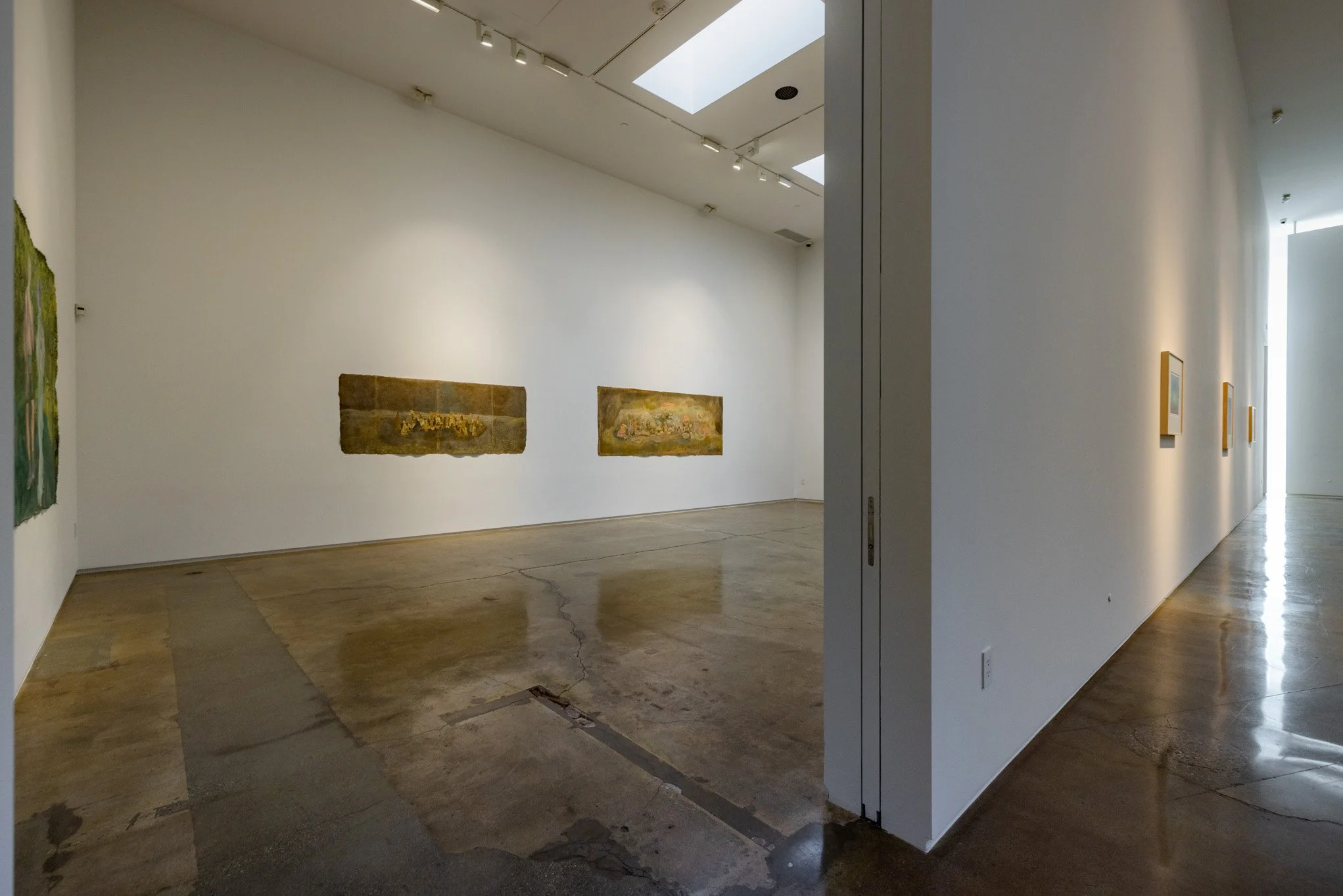 An art gallery with white walls, featuring abstract paintings and large skylights providing natural lighting; polished concrete floors.