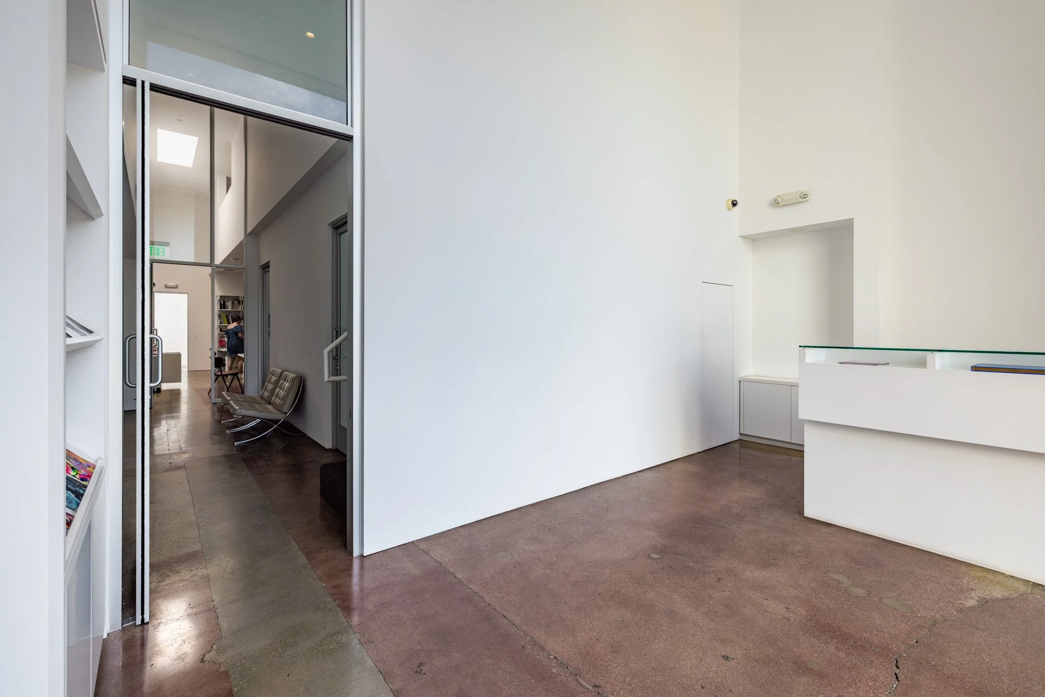 Empty gallery with white walls, brown polished concrete floor, and an open doorway leading to a room with chairs and bookshelves.
