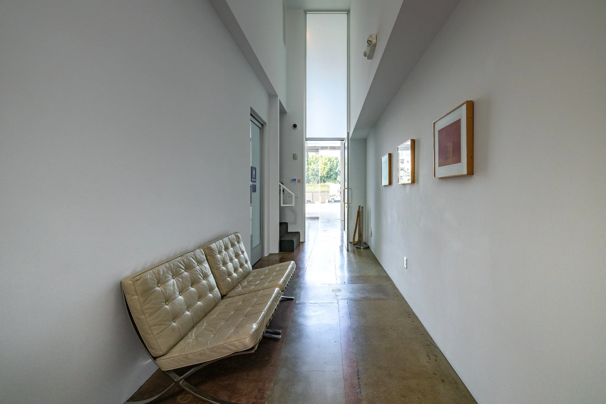 Minimalist empty hallway with three framed pictures on the right wall, beige leather couch on the left, and glass door at the end of the corridor with natural light.
