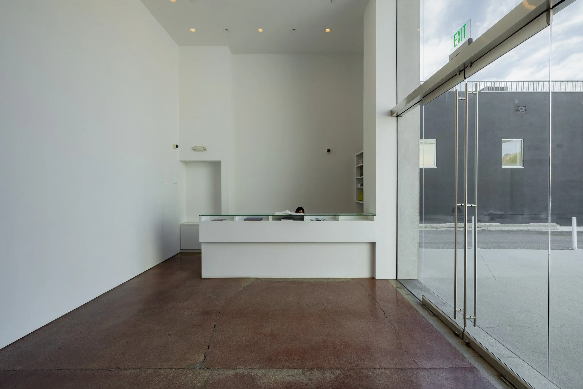 Empty modern art gallery with white walls, brown concrete floor, and a reception desk with a person partially visible behind it. Glass entrance door on the right with an exit sign above.